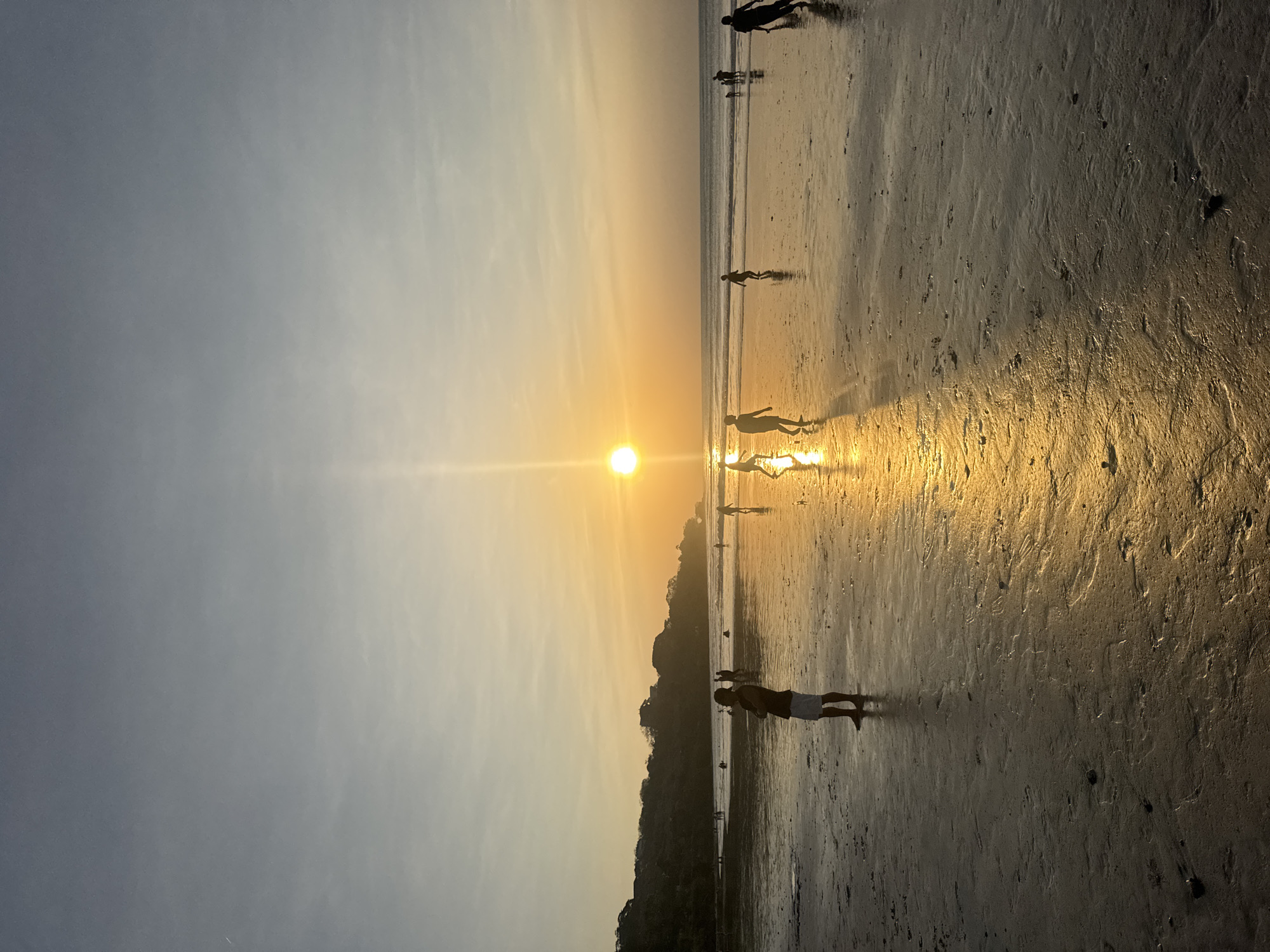 An aerial perspective shot captures a wide expanse of a beach during sunset, casting a warm, golden glow across the scene. The sun, a bright orb, is positioned slightly left of the center, its rays reflecting off the wet sand and creating a shimmering path towards the foreground. The horizon is a blend of soft blues and oranges, transitioning into a clear sky. In the distance, a lush, green hillside is silhouetted against the sky, with structures visible on its crest.
The beach itself is a broad, wet surface, imprinted with the textures of sand and shallow water, reflecting the vibrant sunset. Scattered across the sand are several figures, mostly depicted as silhouettes due to the backlighting. In the mid-ground, a solitary figure stands prominently, facing away from the camera with arms crossed, appearing to observe the sunset. To the left and right of this figure, other individuals are dispersed, some walking, others appearing to engage in activities. In the far distance, near the water's edge, a small cluster of people is visible, some wading or standing in the shallow waves. The overall impression is one of tranquil evening activity on a tropical beach. The location is identified as Kuta Selatan, Indonesia, a region known for its scenic beaches and popular tourist destinations.