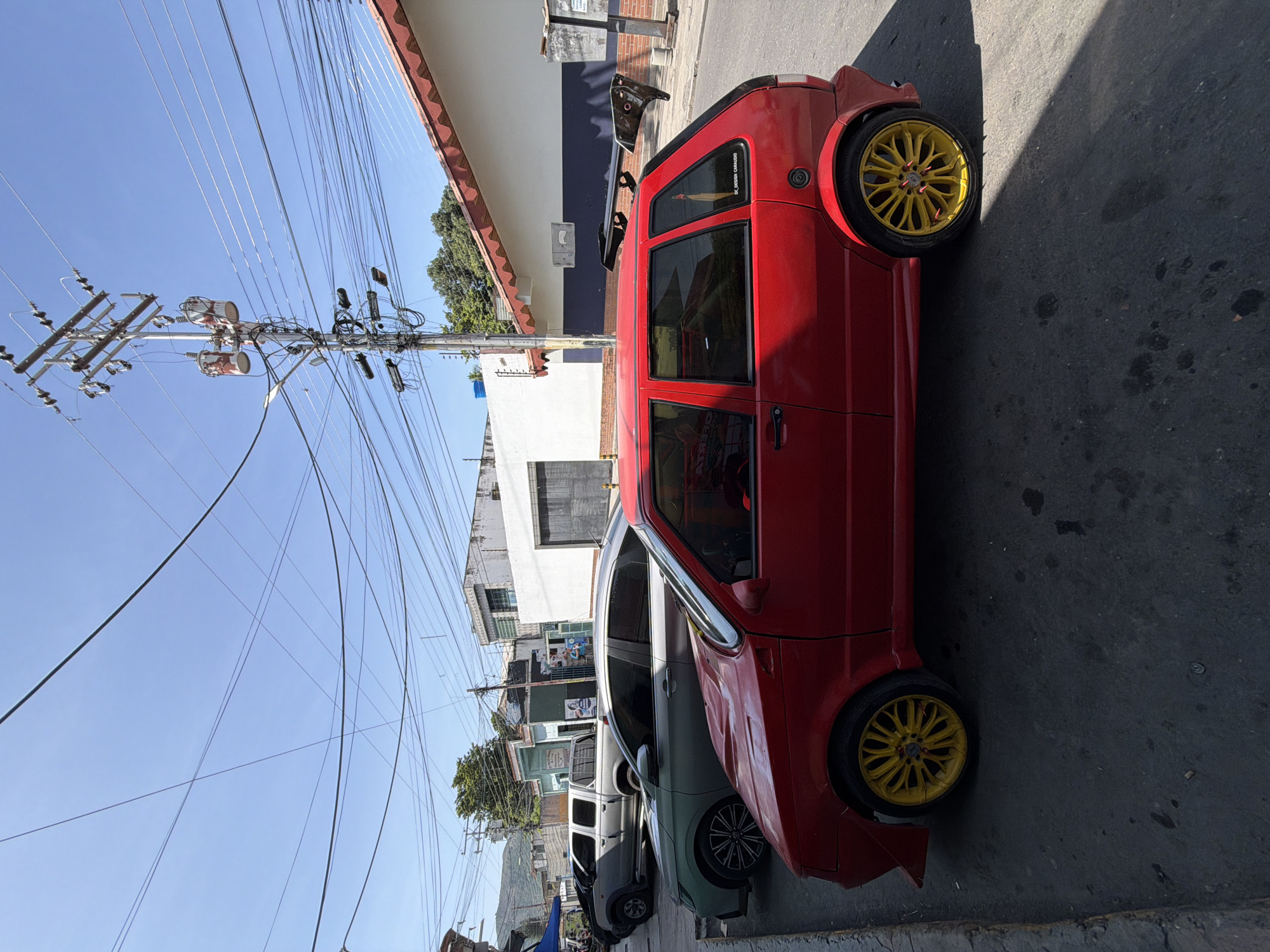 The image captures a street scene in Turmero, Venezuela, under a clear blue sky. In the foreground, a vibrant red hatchback automobile with bright yellow rims is parked on the right side of a paved road. The car features an aftermarket spoiler on its rear. To the left of the red car, a silver sedan is visible, and further behind it, a silver pickup truck is parked. The background reveals a row of buildings with varying architectural styles, including a white building with a red-tiled roof and a white structure with a dark blue stripe. Utility poles with numerous overhead wires dominate the upper portion of the image, suggesting a densely wired urban environment. The overall scene depicts a typical day in a Venezuelan town, with parked vehicles and the infrastructure of a residential or commercial street.