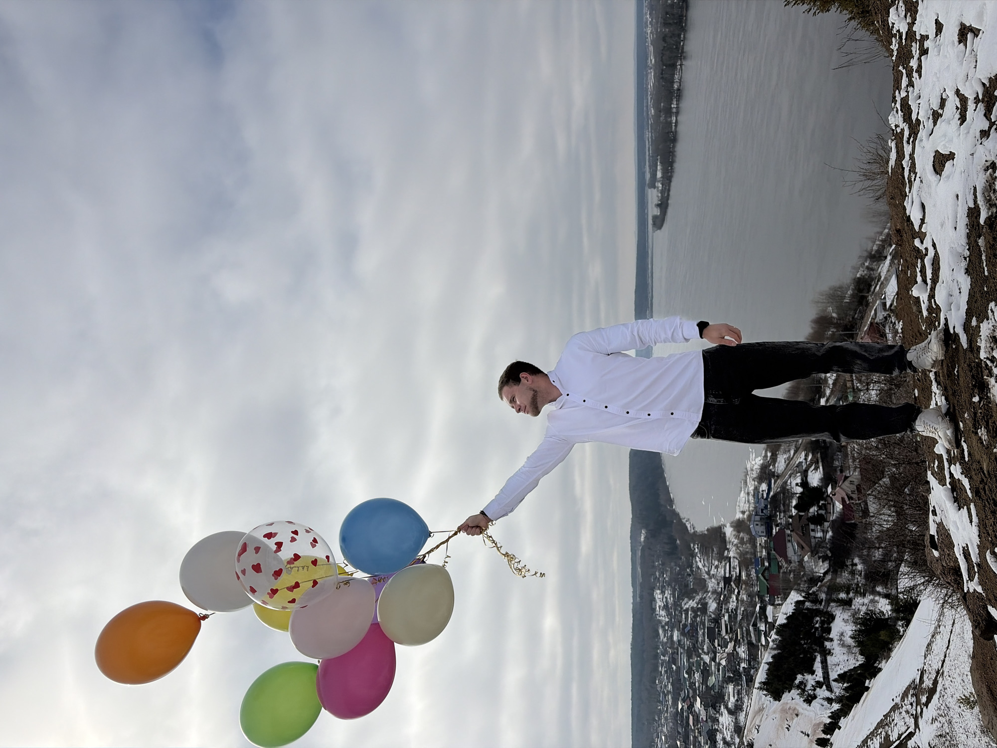 A man in a white shirt and black pants stands on a snowy cliff holding a bunch of colorful balloons. The location is Ольховка, Russia, as indicated by visual cues of the cityscape and landscape visible in the background. A body of water lies beyond the town. The sky is overcast and cloudy.
