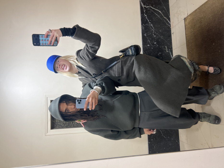Two individuals are positioned within an interior space, likely a hallway or lobby, captured in a mirror selfie. The background consists of a plain light-colored wall, featuring a dark veined marble wainscoting along the base. A light-colored doorframe with a darker recessed panel is visible in the upper left background. The floor is light-toned, with a dark brown rectangular mat positioned in the lower right foreground.
The subject on the left is a male with dark skin and long dreadlocks, which are partially covered by the hood of his dark olive-green oversized sweatshirt. He wears dark grey baggy trousers and dark boots. Accessories include a large silver-toned ring on his left index finger and a green ring on his left ring finger. He holds a light blue smartphone in his right hand, pointed towards the mirror, seemingly initiating the photograph. His left arm is draped over the shoulder of the individual to his right, and his expression is neutral.
The subject on the right is a female with light skin and blonde hair, wearing a dark blue cap. She is dressed in a long, dark grey/brown double-breasted coat over a black long-sleeved top, with patterned socks visible above her black Mary Jane-style shoes, which are embellished with silver studs and feature a strap across the instep. She carries a small black shoulder bag with gold "Miu Miu" lettering, worn across her body. In her raised left hand, she holds a black smartphone encased in a cover featuring small red/pink motifs. Her right leg is slightly bent and lifted. Her left arm rests on the male's shoulder. Her facial expression is exaggerated, characterized by an open mouth revealing teeth and squinted eyes, conveying a playful grimace.
The precise location within the city of Paris, France, is not discernible from the visual information presented, as the scene depicts a generic interior setting.