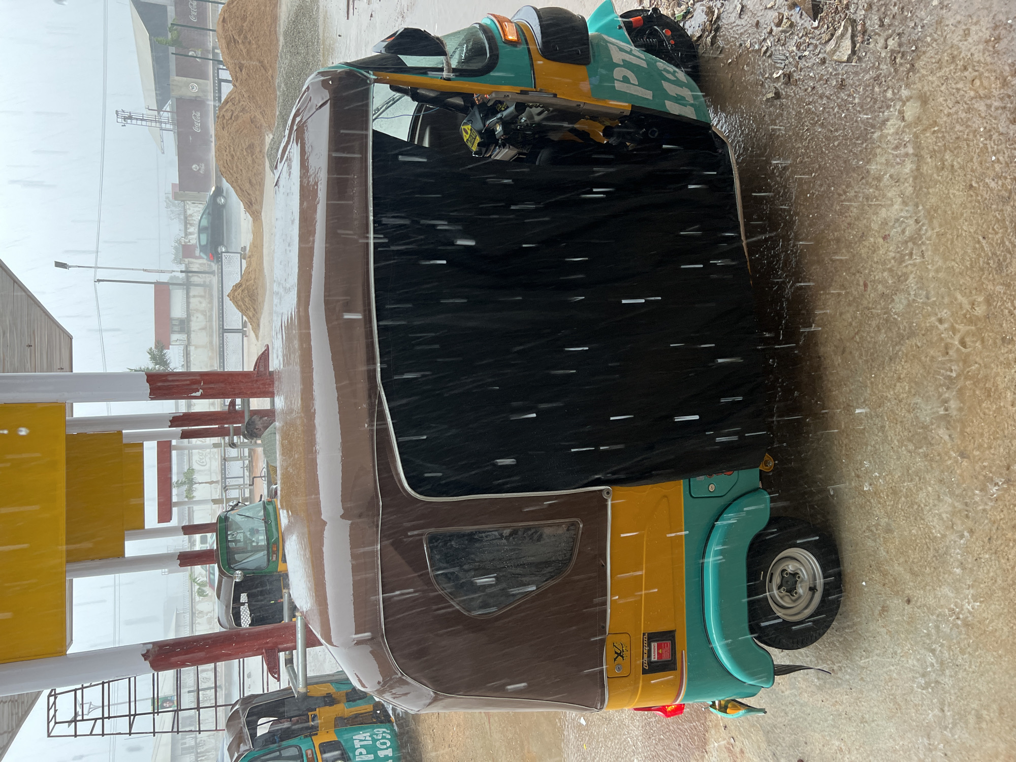 The image shows a yellow and teal auto rickshaw parked on a wet surface, presumably a street, in Barakin Lerawi, Nigeria. It is raining heavily. The rickshaw has a brown canvas top with a black panel covering the front opening. Other rickshaws are visible nearby. In the background, there are piles of sand or gravel, a Coca-Cola branded building, and other structures, all blurred by the rain.