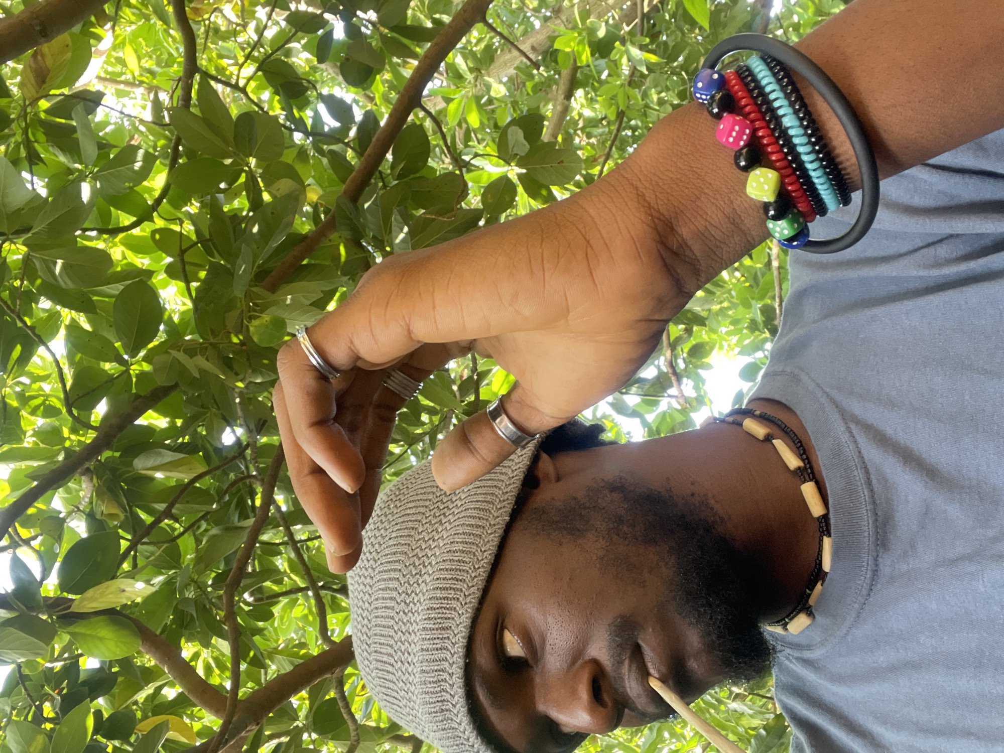 A man with dark skin, a beard, and a gray knit hat is seen from a low angle, looking up into the branches of a tree with green leaves. He has several rings on his fingers and a collection of bracelets on his wrist, including one with colorful dice. He is wearing a gray t-shirt and a necklace with wooden beads. A thin, light-colored object is in his mouth, possibly a straw or a thin stick. The background consists of foliage, with branches and leaves filling the upper portion of the frame. It is not possible to determine the location in the city of Jos, Nigeria from the image alone.
