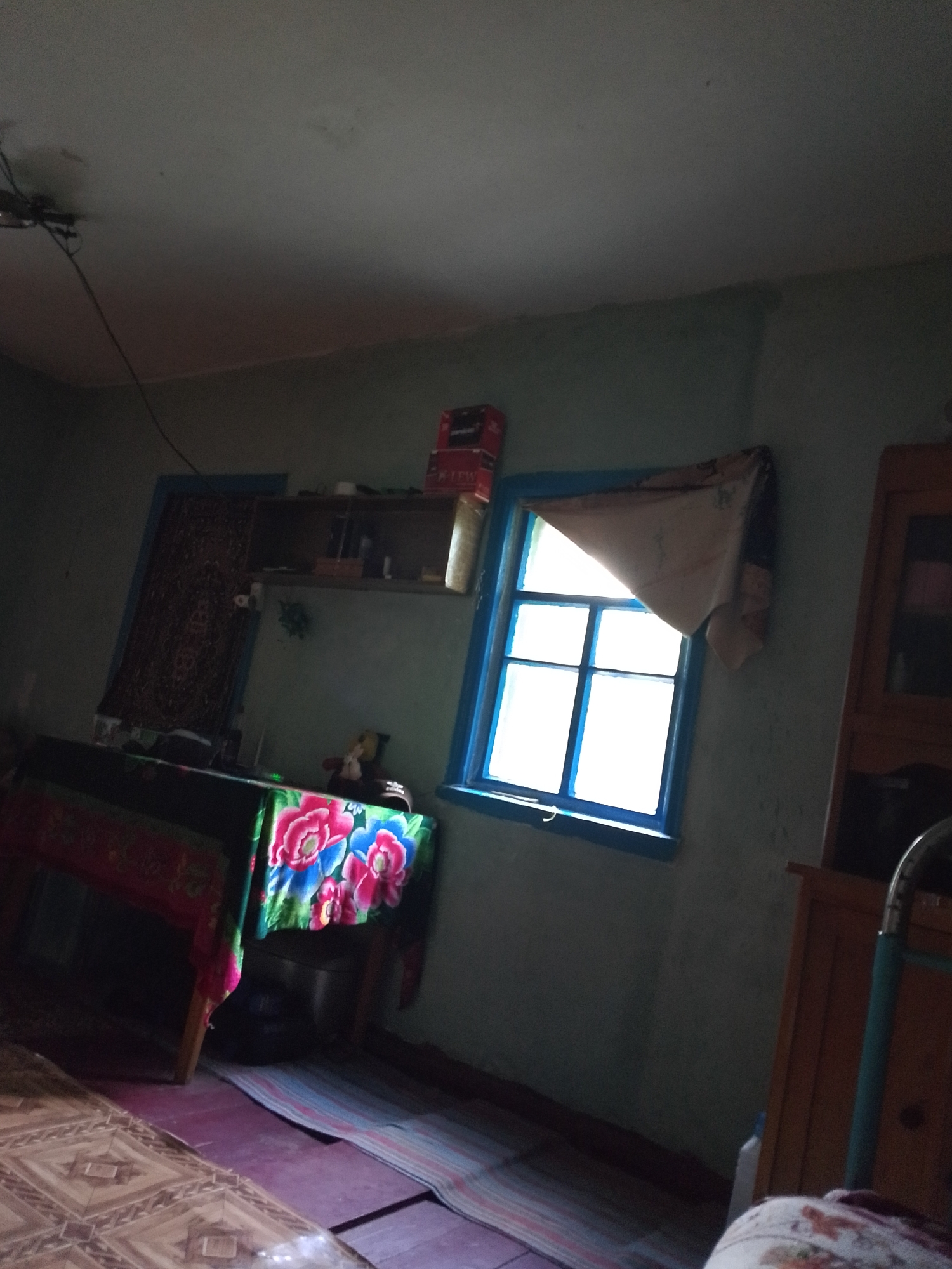 The image depicts the interior of a room with a muted, blue-green wall. A window with a blue frame is positioned on the right side of the frame, allowing bright daylight to enter. The window has a light-colored curtain that is partially drawn. To the left of the window, a wooden cabinet with glass doors is visible, containing some objects. In the foreground, a table is covered with a colorful cloth featuring floral patterns. A rug with horizontal stripes is laid out on the floor. In the background, to the left, there is a tapestry or rug hanging on the wall, and a shelf above it holds several items, including boxes and containers. A ceiling fan is mounted on the ceiling, with a wire extending from it. The overall lighting suggests it is daytime, with light streaming in from the window. The scene appears to be in a residential setting.