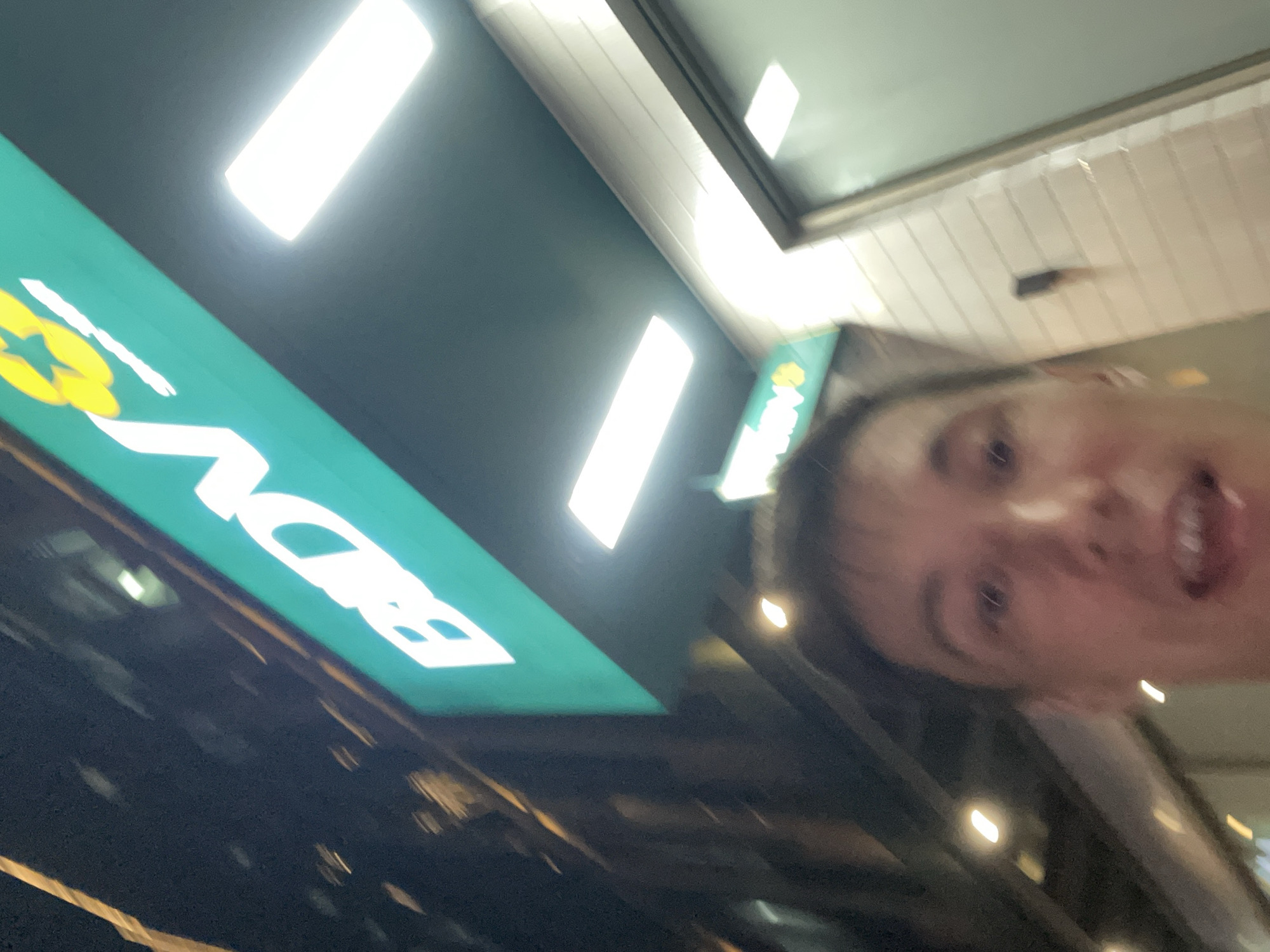 A selfie taken at night in Thủ Đức, Vietnam, shows a person's face from the chest up in the foreground. They are looking at the camera with their mouth slightly open. Behind them, a brightly lit green sign with white text that reads "BIDV" and a yellow logo is visible, indicating the presence of a bank. The sign is mounted on a dark-colored overhang. In the background, there are glimpses of a brick-patterned wall and a glass door, typical of a commercial building. The overall lighting suggests an urban setting at night, with streetlights and building lights illuminating the scene. The image is slightly blurred, suggesting movement during the photograph's capture.