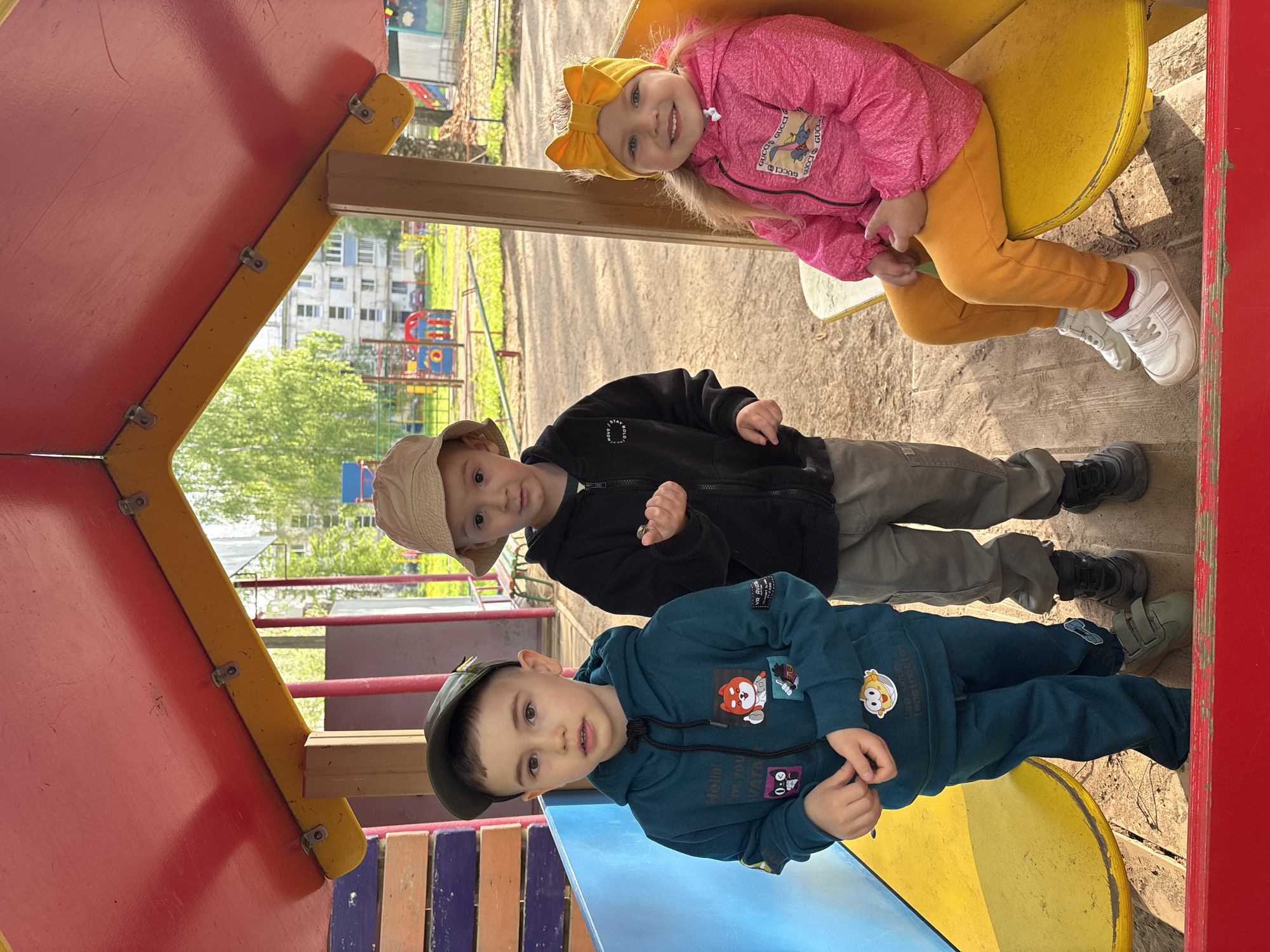 The photograph shows three children on a brightly colored playground structure. A young girl is seated at the top, wearing a pink jacket, yellow pants, white sneakers, and a yellow headband with a bow. Below her is a boy in a black zip-up jacket, grey pants, and a light-colored hat. At the bottom of the frame, another boy wears a blue hooded sweatshirt, matching pants, a dark baseball cap, and green sneakers. They are looking at the camera. The playground has red, yellow, and blue components. In the background, a multi-story residential building, trees, and other playground equipment are visible. The image appears to have been taken outdoors on a sunny day. The picture was taken in Chaikovsky, Russia.