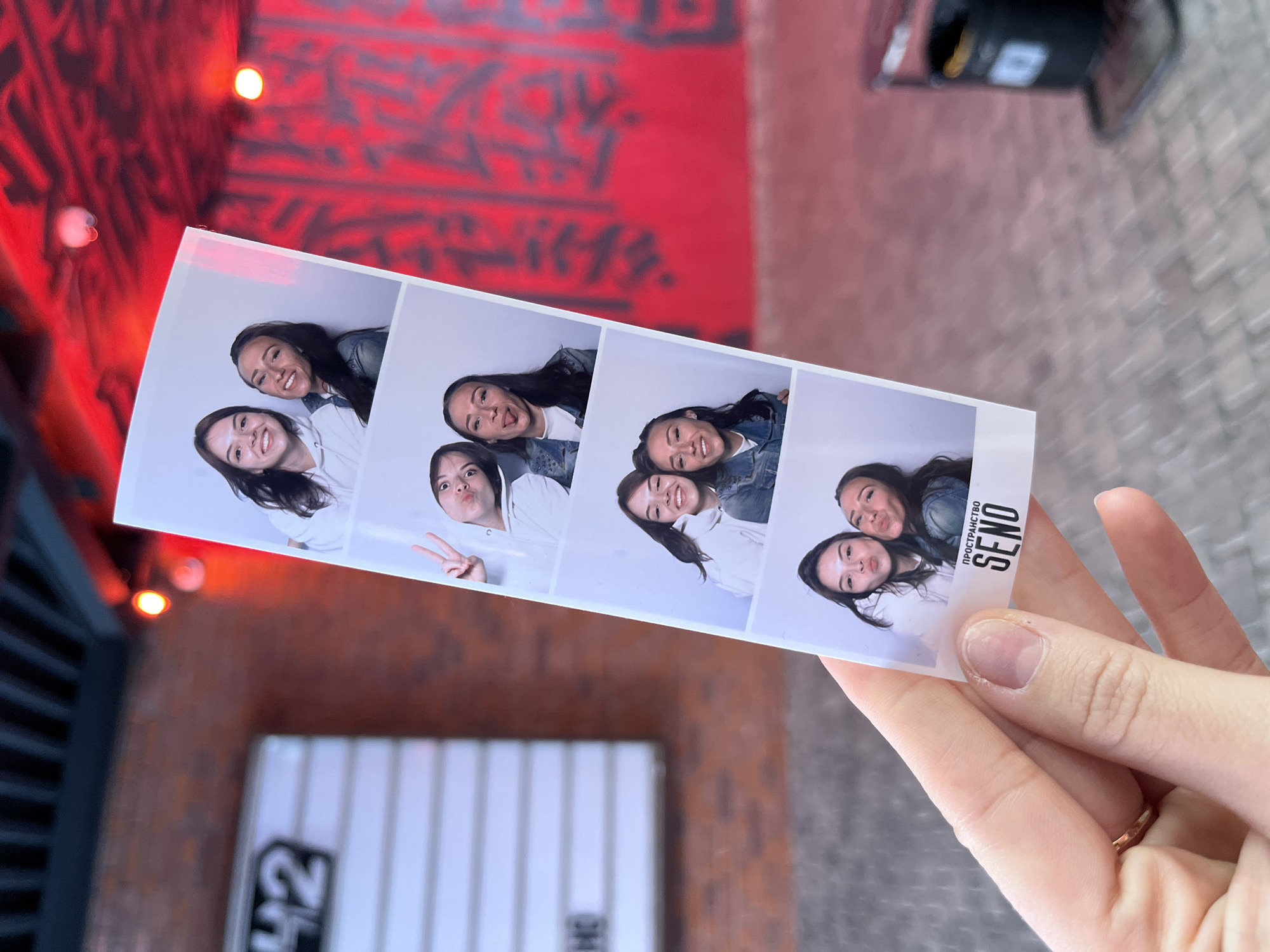 In Saint Petersburg, Russia, a person's hand is holding a photo booth strip featuring four frames of two young women posing. The background is a brick wall with a sign reading "42" and a bright red patterned wall with a trash can. The photo strip shows the women making various playful faces, with one of them sticking out her tongue and another giving a peace sign. The bottom frame includes the words "ПРОСТРАНСТВО SEND". The scene appears to be outdoors, possibly at a venue or event.