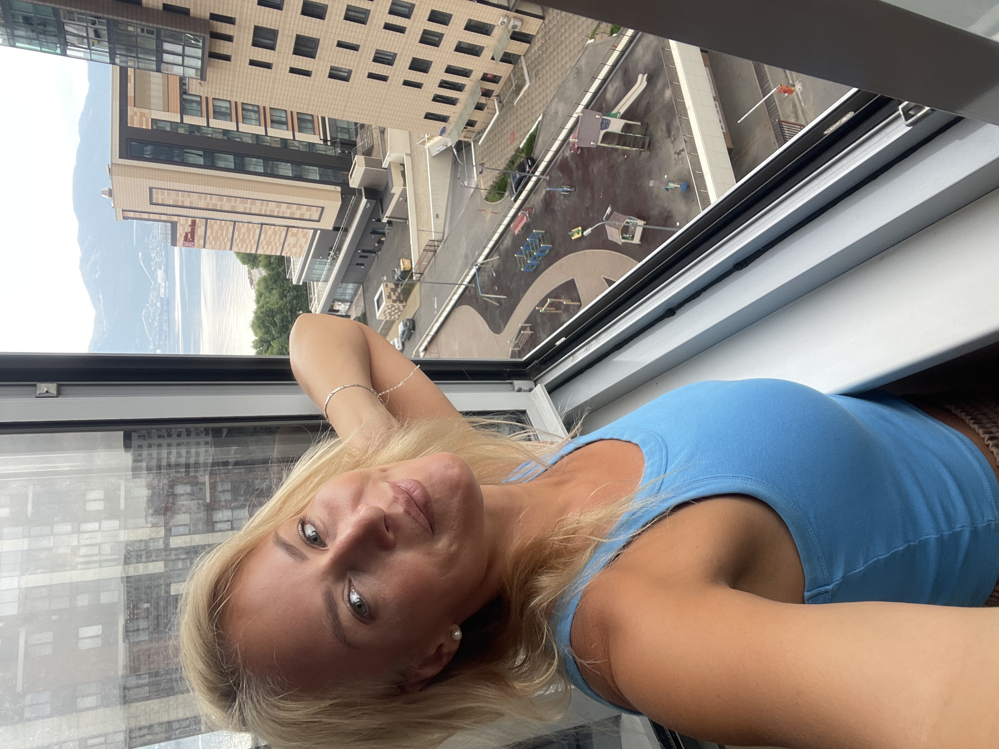 An outdoor, eye-level, medium shot captures a woman with blonde hair in her late 20s or early 30s posing for a selfie by a window. She is wearing a blue tank top and a bracelet on her right wrist. Her left arm is bent upwards, with her hand resting on her head. The background reveals a view of a city, likely Novorossiysk, Russia, with modern buildings, a body of water, and mountains in the distance. Below the buildings, a playground area with slides and swings is visible. The overall lighting suggests an overcast day.