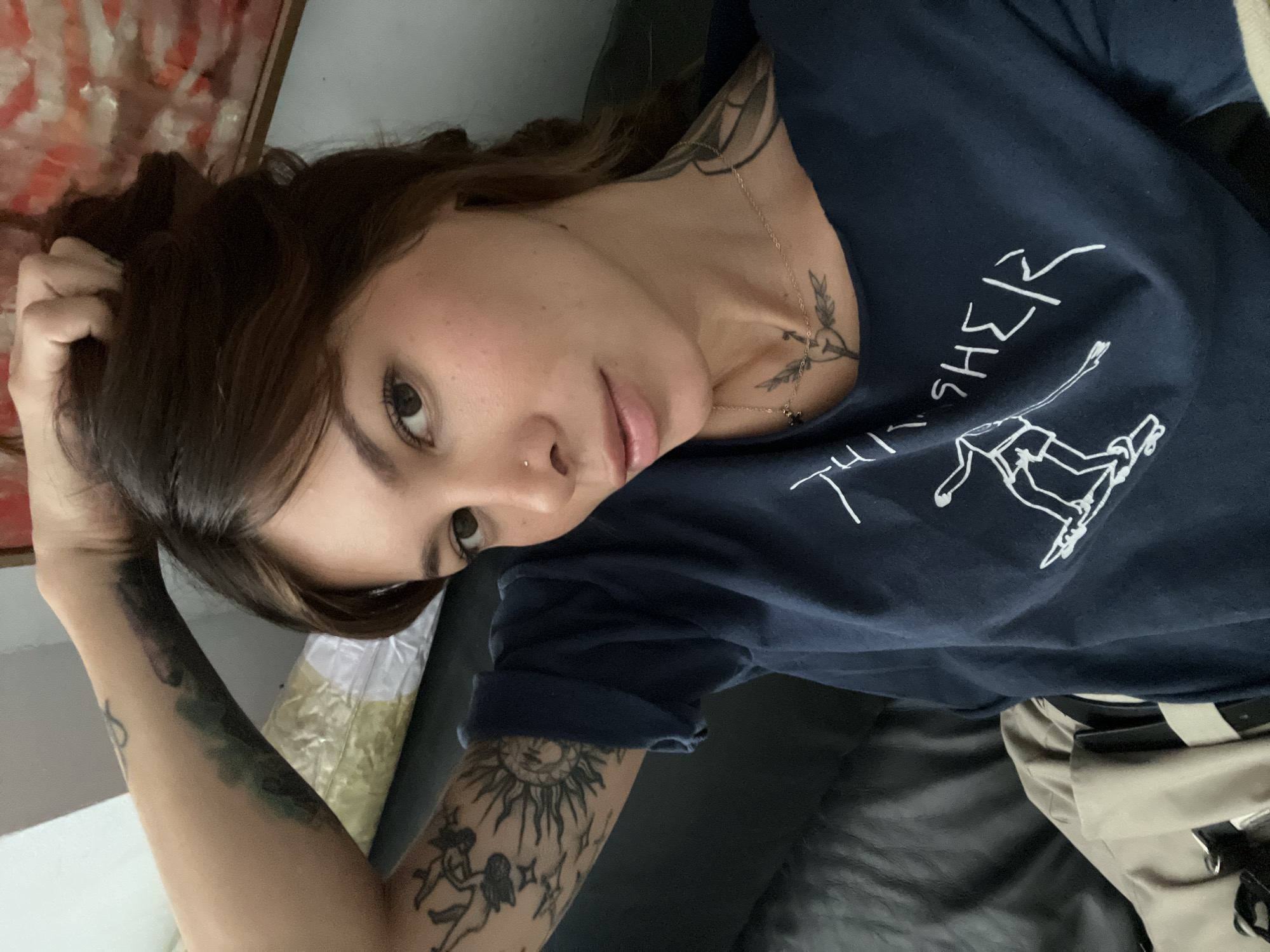 The image is a close-up, selfie-style shot of a woman with brown hair, wearing a dark blue t-shirt with white graphics. She has visible tattoos on her left arm and neck, and a nose ring. Her left arm is raised, with her hand in her hair. She is lying down, likely on a black leather surface, with a beige bag visible in the lower left of the frame. The background is out of focus, with hints of a painting with red and white tones on the right. The lighting suggests an indoor environment. No location in Barcelona, Spain is identifiable from the image.