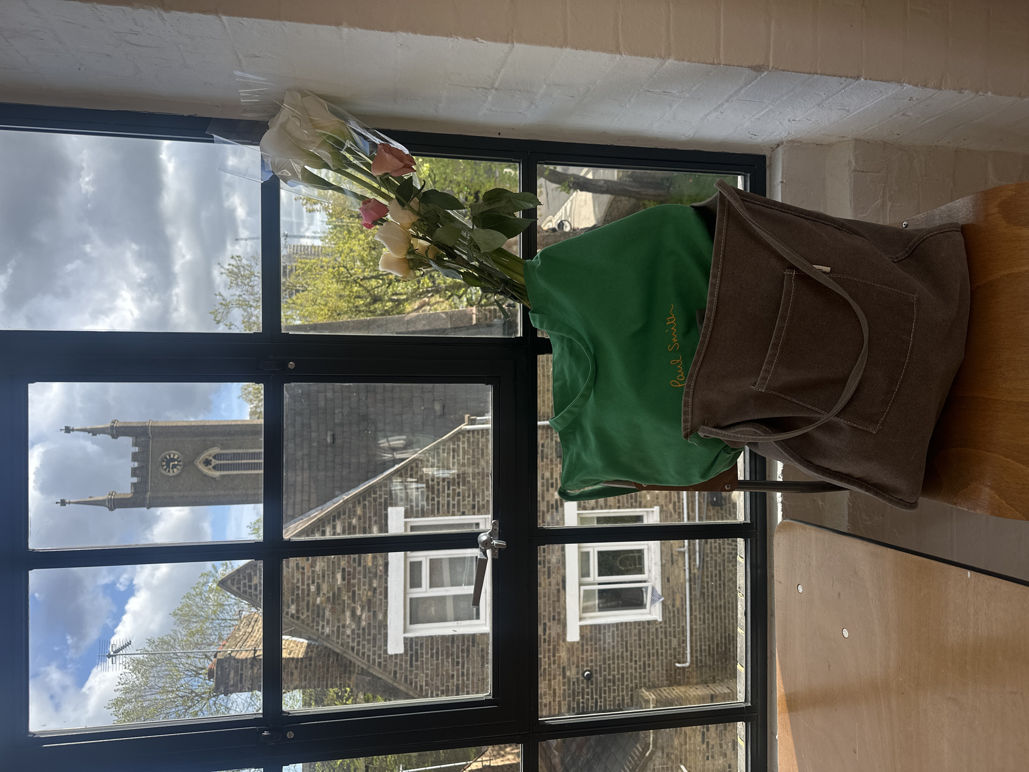 The image presents an interior view looking out through a large, black-framed window. On the right side, a light-colored wooden surface holds a brown canvas tote bag. Next to the bag, a bright green sweatshirt or t-shirt with "Paul Smith" embroidered in yellow script is visible. To the left, a bouquet of roses, primarily white and light pink/yellow, is wrapped in clear plastic. Above the window, a textured white wall, possibly painted brick, is seen.
Through the window, a cloudy daytime sky is visible in the upper left. A prominent grey stone church tower with a clock face and spires occupies the upper central panes. Below and to the right of the church, various brick residential buildings with white-framed windows and rooftops featuring an antenna are visible. Trees with green foliage indicate a spring or summer setting.
No people or specific activities are depicted; the scene is static. The architectural style of the church and brick buildings, combined with the provided context, suggests a location in London, United Kingdom. The time of day is daytime, with bright but cloudy weather.
The only visible text is "Paul Smith" on the green garment.