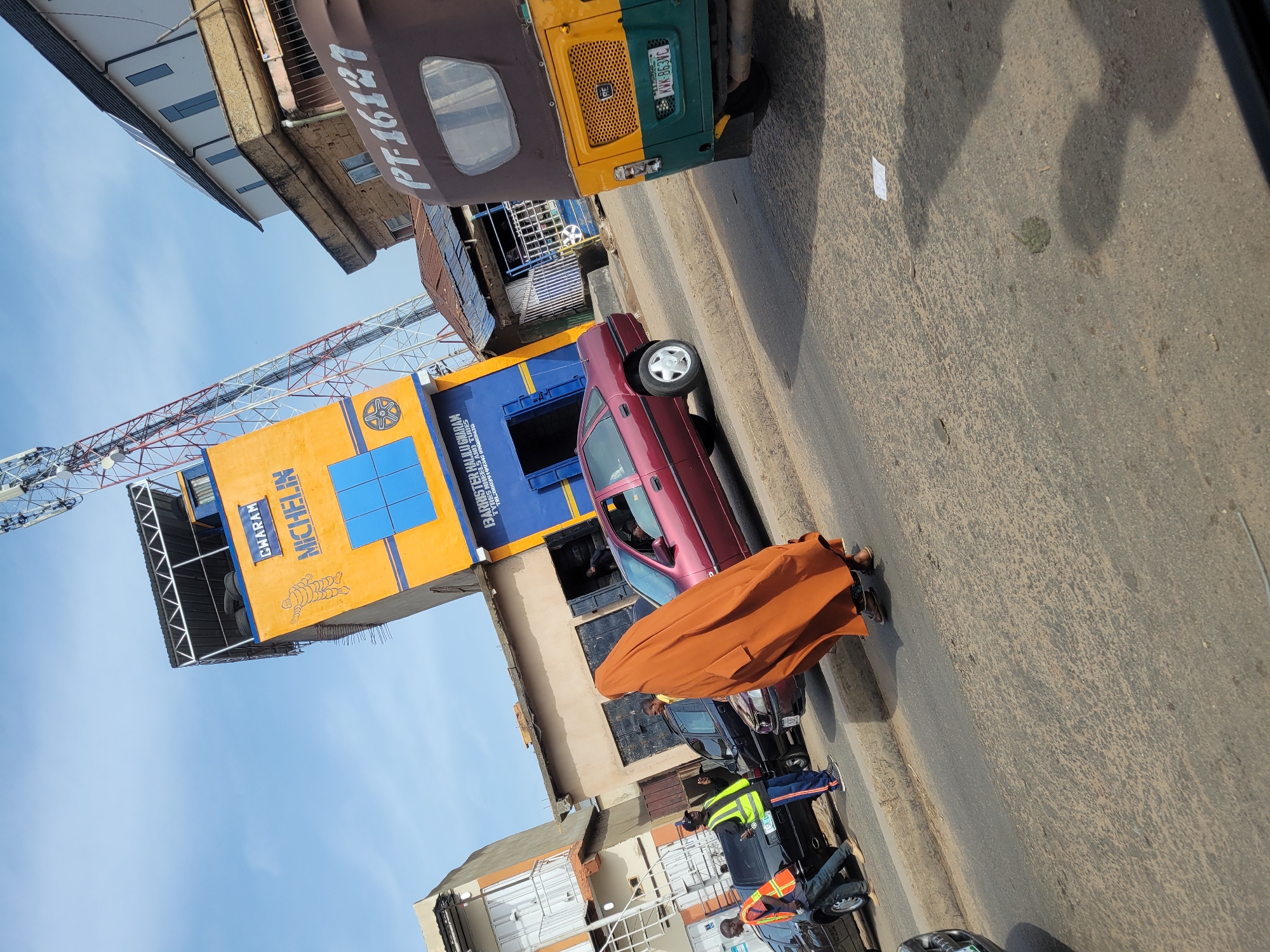 The image captures a street scene in what appears to be a city in Unknown, Unknown. A pedestrian wearing a large, brown, cloak-like garment walks on the street next to a maroon car. Nearby are road workers or traffic wardens wearing reflective vests and pants. Cars are also visible. In the background, there is a tall building with a large, rectangular Michelin advertisement painted on its side. A yellow and green auto-rickshaw (tuk-tuk) is parked on the side of the road. Overhead, there are metal structures, possibly part of a bridge or tower. The sky is visible in the background, suggesting it's daytime. The road surface is dark and appears to be asphalt.