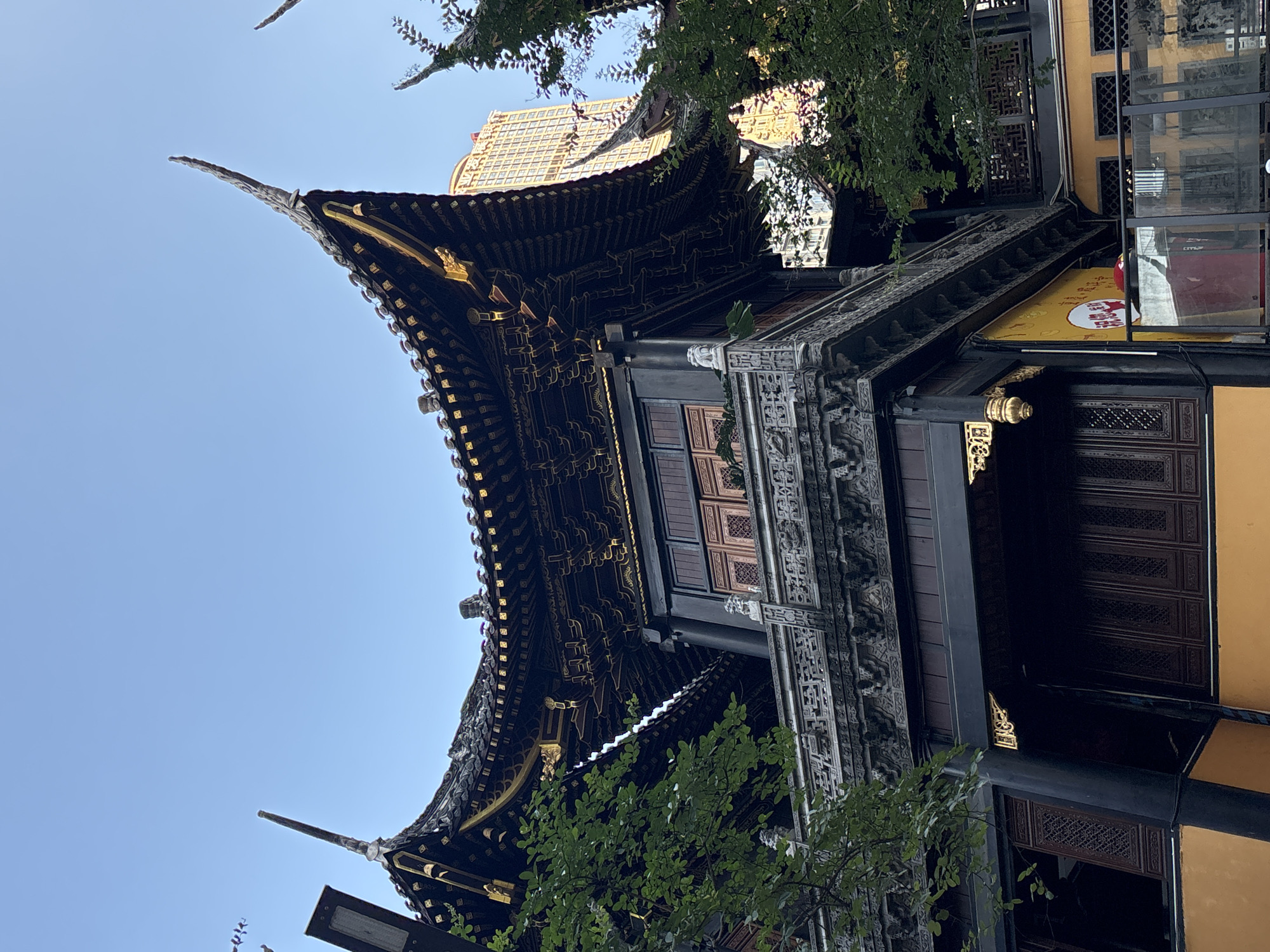 The image shows a traditional Chinese temple building with ornate, dark wooden architecture and a curved, tiled roof adorned with gold accents. The building is viewed from a low angle, looking upwards, with a clear blue sky visible above. Lush green tree branches partially obscure the upper parts of the structure, suggesting it is daytime and the weather is pleasant. The intricate carvings and decorative elements on the building, including what appear to be dragons or mythical creatures, highlight its cultural significance and historical aesthetic. Adjacent to the temple, a more modern building with a yellow facade and glass windows is visible, hinting at a blend of old and new in the surrounding urban environment. A yellow sign with red text and a circular logo is partially visible on the modern building, though the text is not legible. The overall impression is one of a vibrant cultural setting in a district like Yuzhong, China, where ancient traditions coexist with contemporary life.