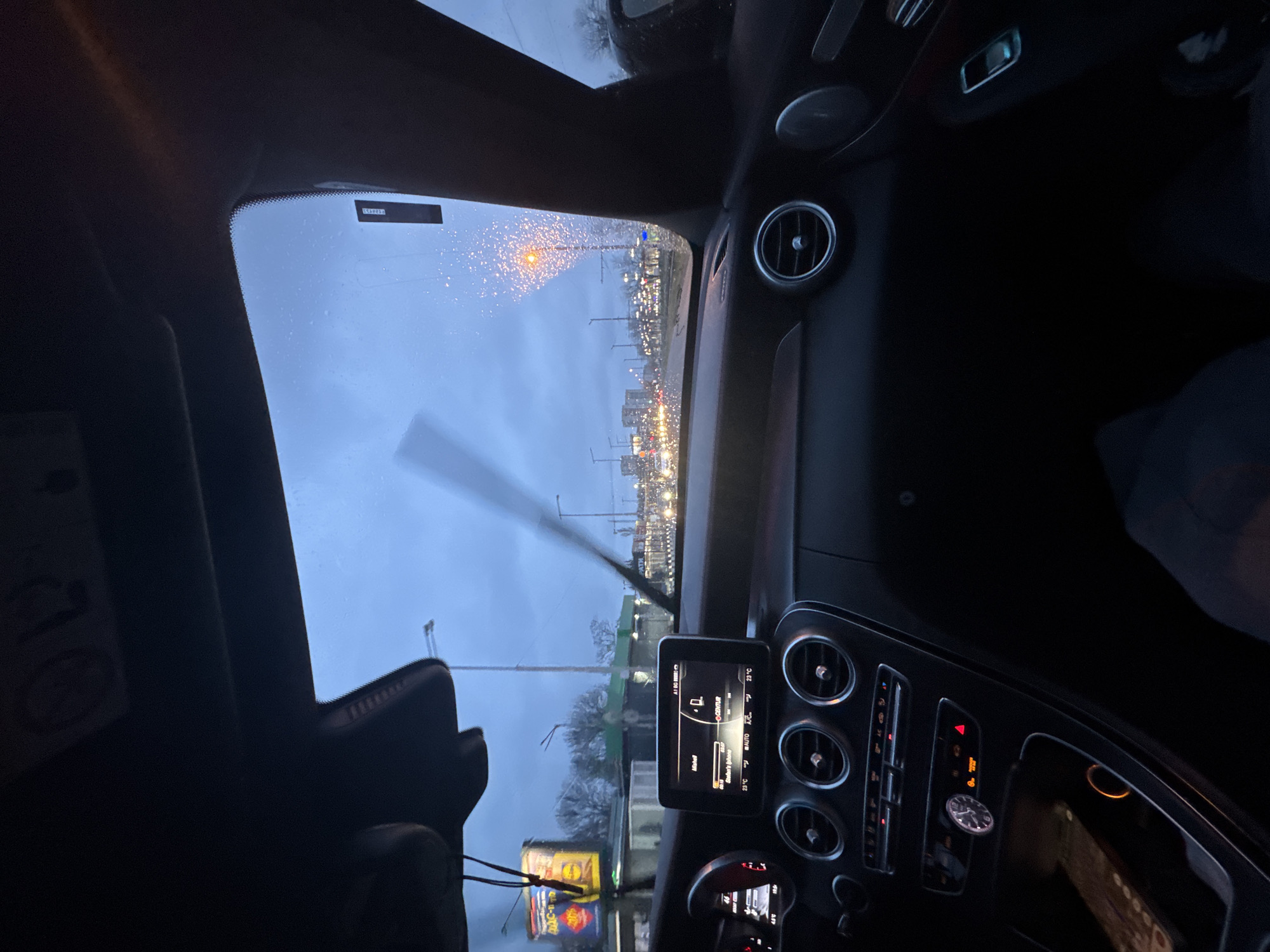 Captured from inside a car during likely rainy weather, the image shows a windshield with raindrops and a blurred view of an urban landscape, presumably in Sofia, Bulgaria. A windshield wiper is visible. Outside, a row of streetlights stretches into the distance, suggesting a main road or boulevard lined with buildings. The dashboard of the car is visible, featuring air vents, a digital display indicating weather conditions, and various buttons and controls. A Lidl bag is sitting in the passenger footwell. The overall scene suggests traffic or a moment of pause in an urban environment.