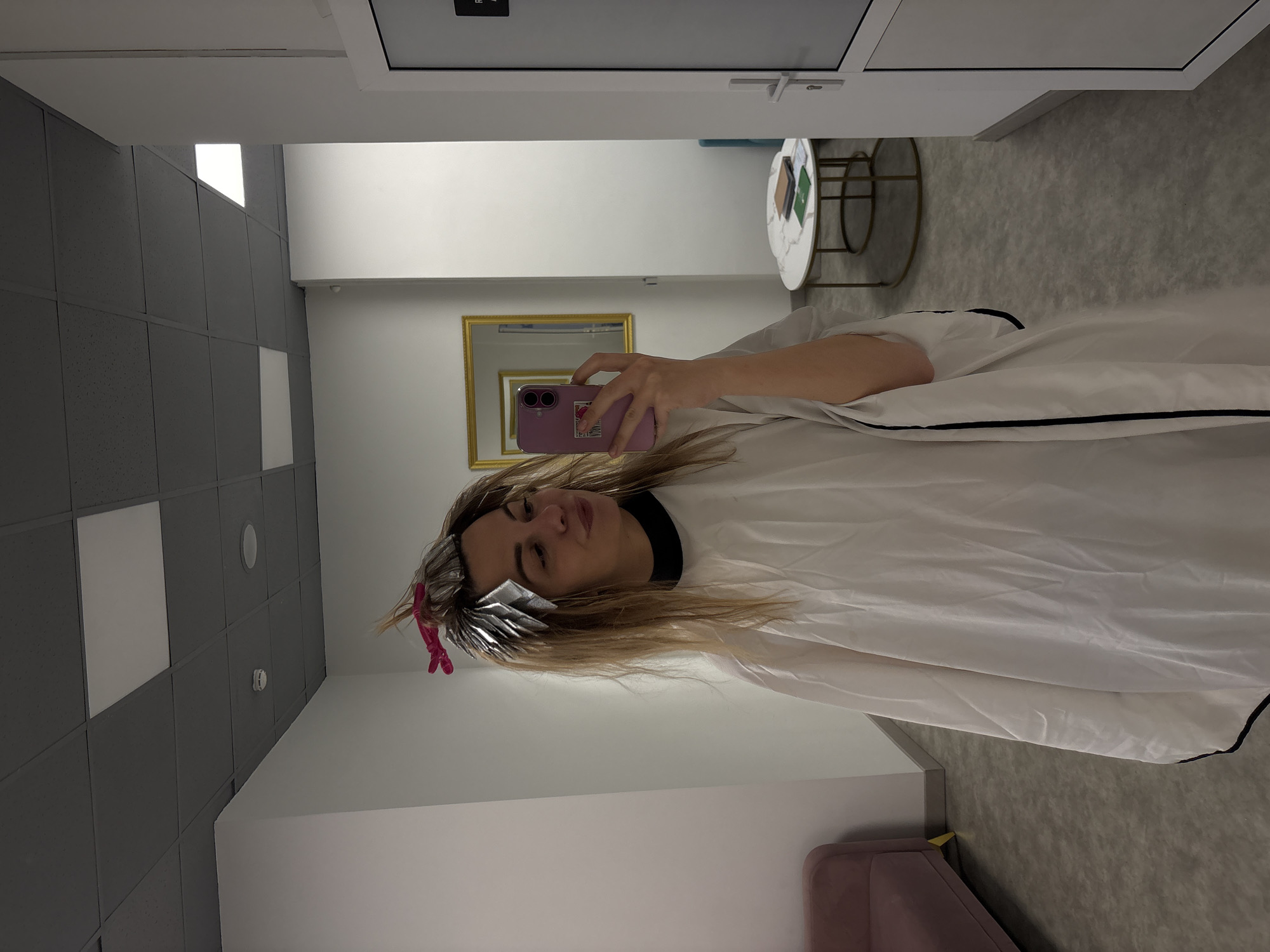 A woman wearing a white salon cape with a black trim is taking a selfie in what appears to be a hair salon. Her hair is being highlighted or dyed, as evidenced by the foil in her hair secured with a pink clip. She is holding a pink smartphone in her right hand. Behind her, there is a mirror reflecting the room and the door. Further in the background, there's a small round table with a white top and a gold frame. The interior of the room is modern, with a gray and white color scheme. The location is in Krasnoyarsk, Russia.