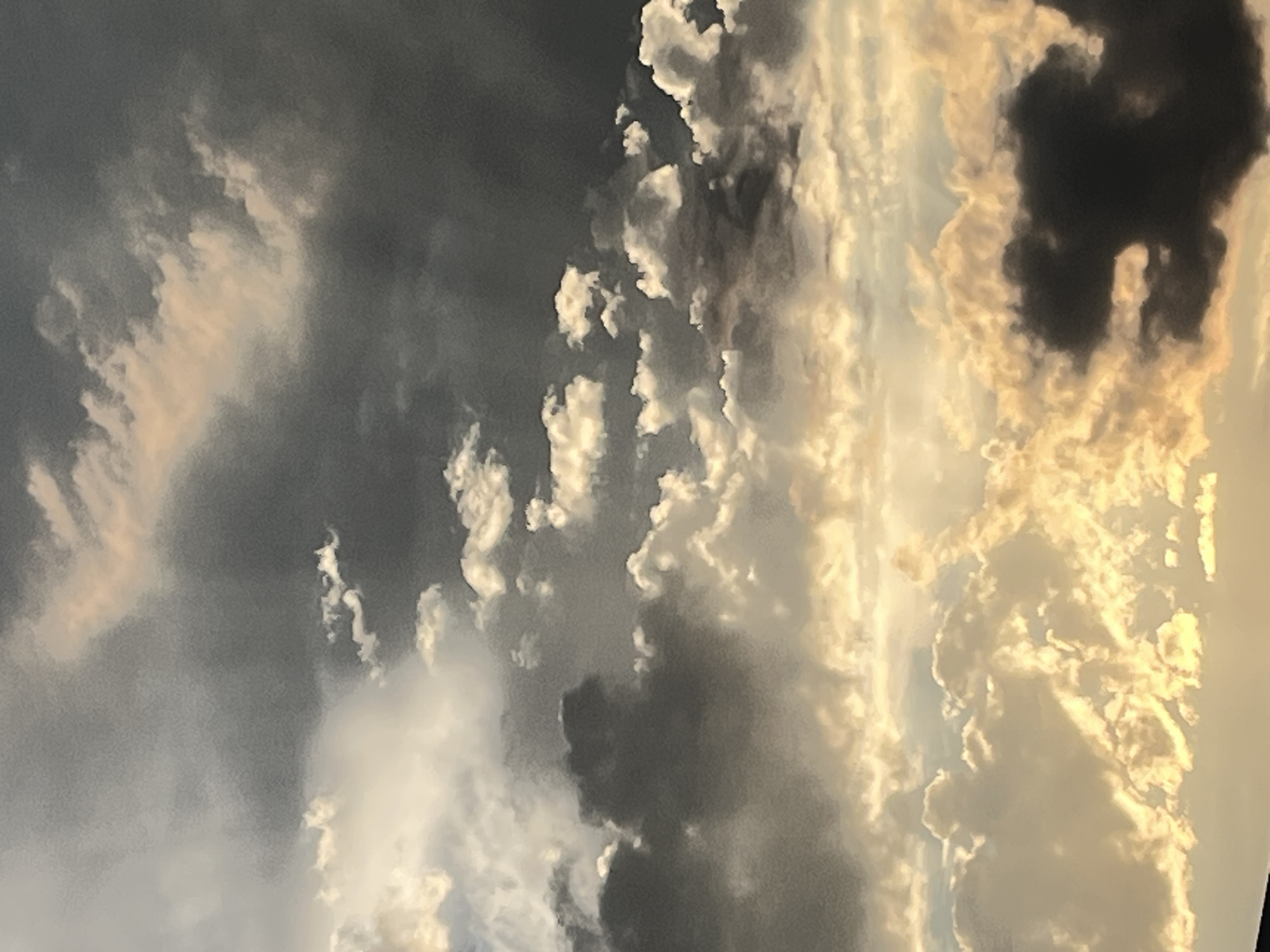 The image is a vertical, close-up photograph of clouds. The clouds are depicted in various shades of gray, white, and golden yellow, suggesting a sunset or sunrise. Some clouds are dark and ominous, while others are illuminated by the sun, creating a dramatic contrast. The scene is abstract and focuses entirely on the textures and light play within the cloud formations. There is no visible text, people, or objects. The overall mood is dramatic and awe-inspiring, characteristic of a dynamic sky. The location context of Krasnodar, Russia, provides a geographic setting for this natural phenomenon.