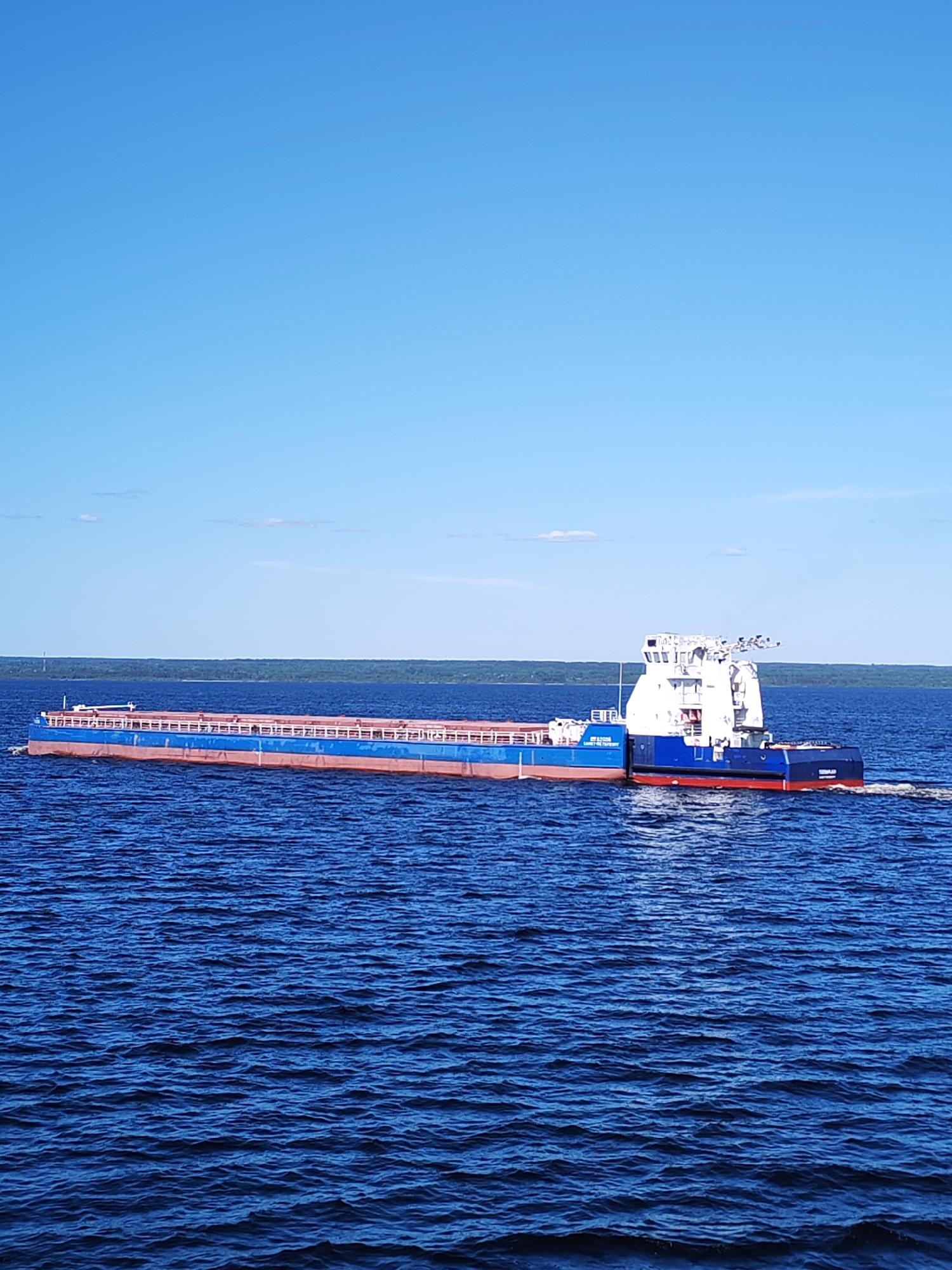 An outbound tugboat, identified by "КОЖУХОВ Л. И." (KOZHUKHOV L. I.) on its white superstructure and "ВОЛГО-ДОН" (VOLGO-DON) on its blue hull, pushes a blue and red cargo barge across a wide expanse of dark blue, rippled water. The vessel moves from left to right, leaving a distinct white wake behind its stern. The barge features a long, open cargo area with white railings. In the background, a distant, dark green forested landmass forms the horizon under a clear, bright blue sky with a few wispy white clouds. The scene is illuminated by natural daylight. No individuals are visible. The location is Unknown, Unknown.
