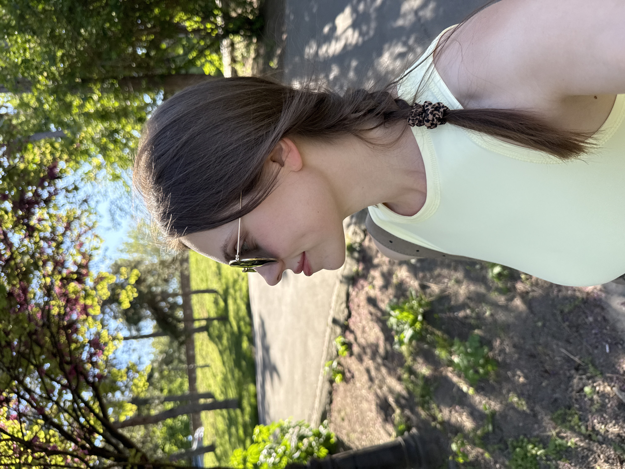 A young woman stands outside on a sunny day. She has brown hair styled in a braid secured with a patterned scrunchie. She wears sunglasses and a light yellow top. The background reveals a sidewalk and grassy area with trees, some of which are flowering. The location is likely a park or green space within Belgrade, Serbia, based on the overall environment and vegetation.