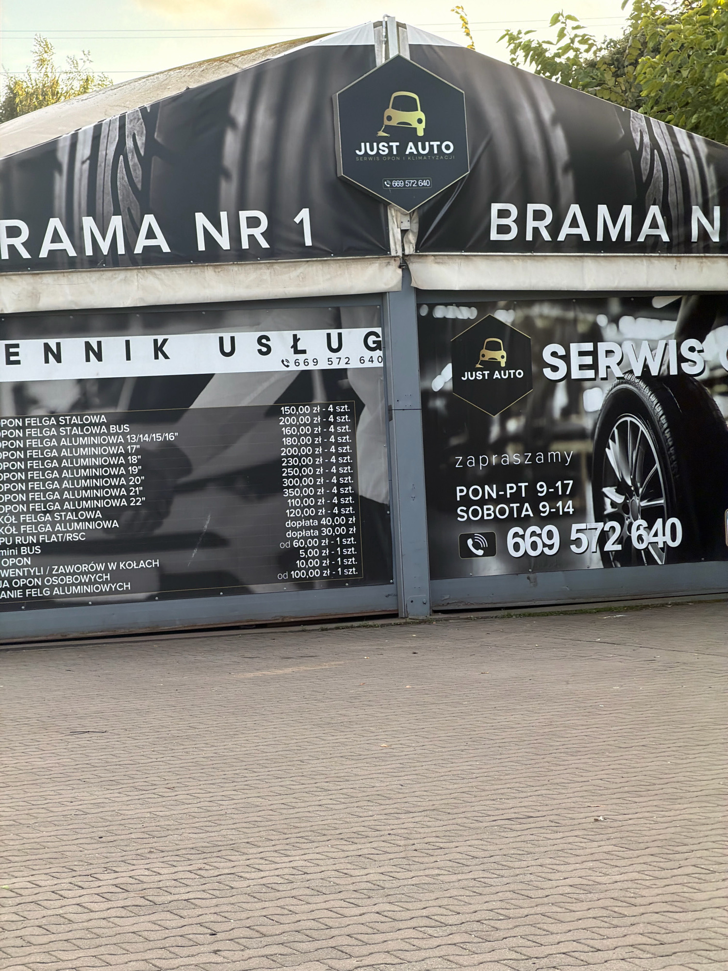 The image shows the facade of a car service establishment called "JUST AUTO" in Wrocław, Poland. The facade features large banners with text and graphics. One banner reads "BRAMA NR 1" (Gate No. 1) and another displays "SERWIS" (Service) alongside an image of a car tire. Below the "SERWIS" banner, operating hours are listed as "PON-PT 9-17" (Monday-Friday 9 AM to 5 PM) and "SOBOTA 9-14" (Saturday 9 AM to 2 PM). A phone number, "669 572 640", is prominently displayed. The lower portion of the facade shows a price list for various tire and wheel services. The ground in front of the establishment is paved with interlocking concrete blocks. The overall scene depicts a commercial establishment offering automotive services.
