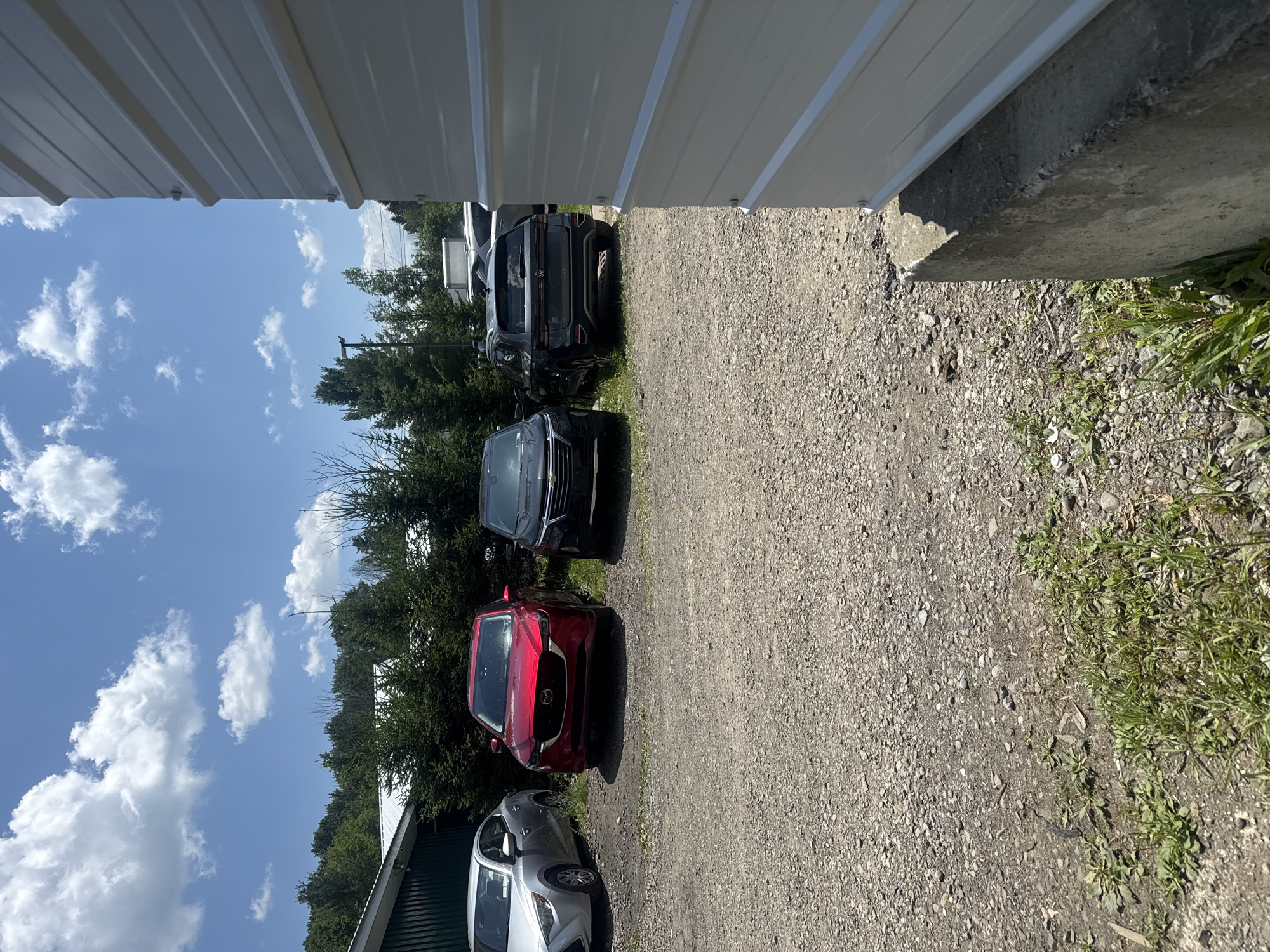 A daytime exterior shot captures a gravel parking lot with three cars parked in front of a dense line of evergreen trees. To the left, a silver sedan is partially visible. In the center, a red SUV is parked. To the right, a dark-colored SUV is parked. A white, corrugated metal building runs along the right edge of the frame, obscuring part of the scene. The sky is blue with scattered white clouds. The scene appears to be located in the Village of Johnson City, United States of America, possibly a commercial or industrial area, given the presence of the building and the arrangement of vehicles.