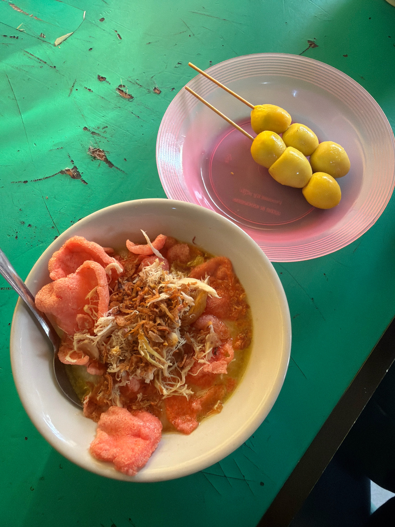 A top-down, medium shot shows two plates of food on a bright green, weathered table. The plate on the left is a white bowl filled with a yellow broth, topped with shredded chicken, fried onions, and bright pink crackers. A silver spoon rests in the bowl. The plate on the right is a light pink, ridged dish holding six yellow quail eggs on two wooden skewers. The table surface is chipped and scratched, with some darker areas suggesting wear and tear. A dark shadow is visible on the bottom right of the frame. The overall scene appears to be a casual meal setting.