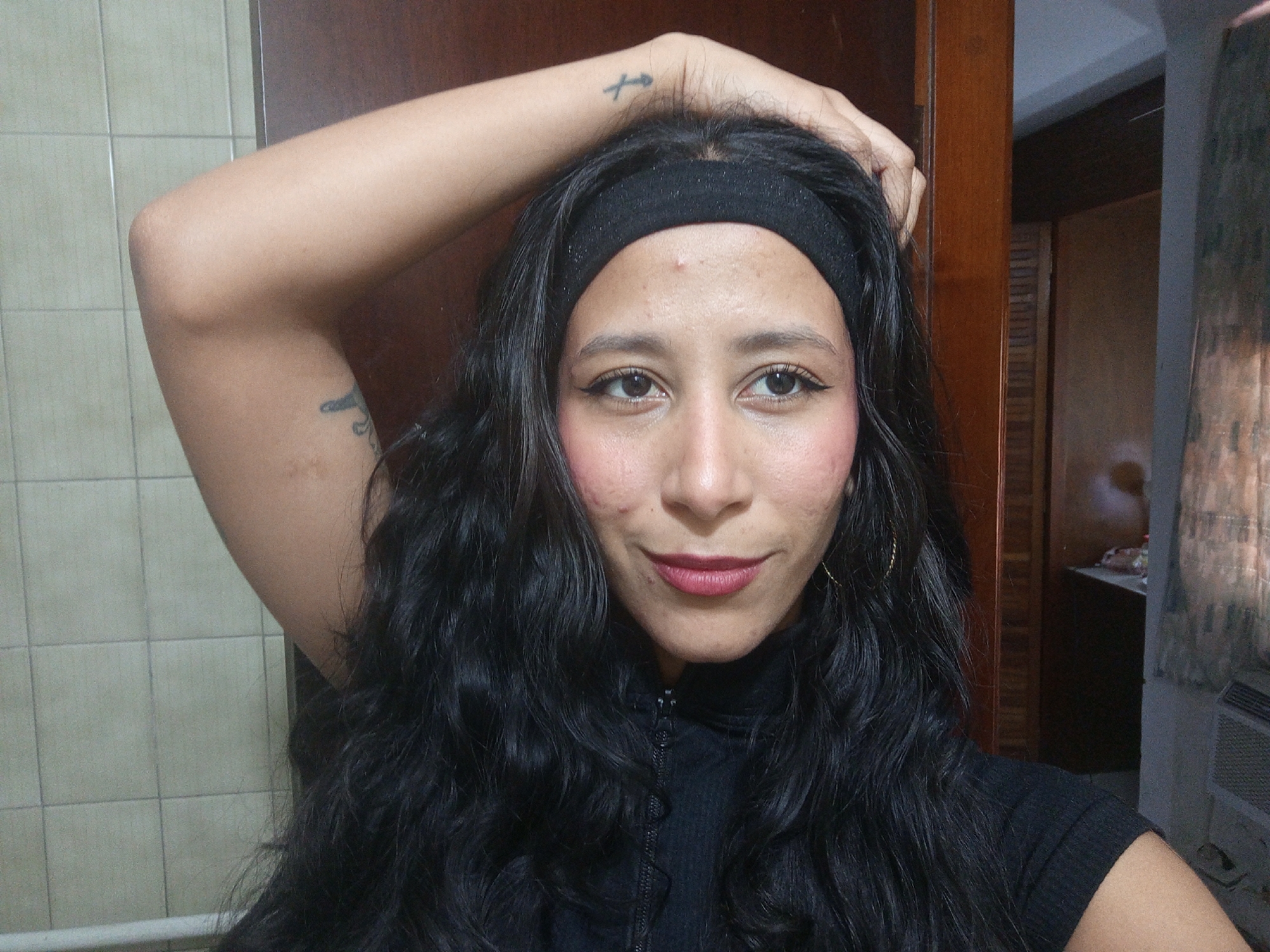 This is a close-up, indoor portrait of a woman in Porlamar, Venezuela. The subject is a woman with long, dark, wavy hair, wearing a black headband and a black ribbed top with a zipper. She poses with her right arm raised, hand resting on the top of her head. She looks directly at the camera with a calm, confident expression and a slight smile. Her dark eyes are accentuated with eyeliner, and her lips are colored with a dark pink or berry-toned lipstick. Rosy blush is visible on her cheeks, and there are some visible blemishes or skin texture on her forehead and cheeks.
Notable details include a small black tattoo on her right wrist, which appears to be a stylized Pisces symbol. Another partial, indistinct tattoo is visible on her left upper arm. A gold hoop earring hangs from her right ear. The background on the left features a light-colored tiled wall, while the right side shows a wooden door or piece of furniture. The lighting suggests the photograph was taken during the daytime.