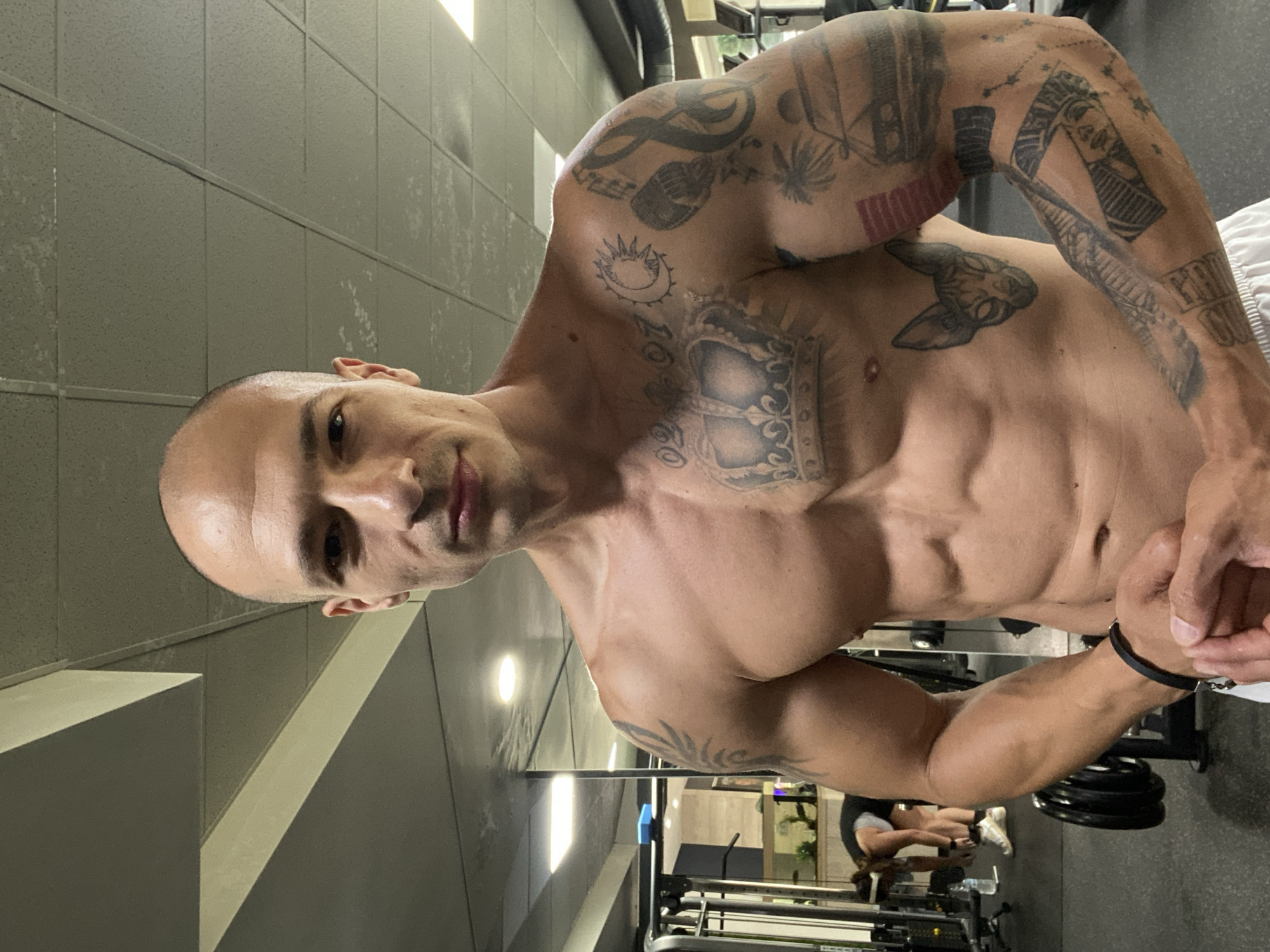 A bald, muscular man with numerous tattoos poses for a selfie in a gym. He is shirtless, wearing white shorts. His tattoos include a crown on his chest, a cat's face, a sun, a microphone, a musical note, palm trees, a red rectangle, and various other designs on his arms and chest. In the background, a woman is seen bending over, possibly tying her shoes. The gym environment features weightlifting equipment, mirrors, and overhead lighting. The image appears to be taken indoors.