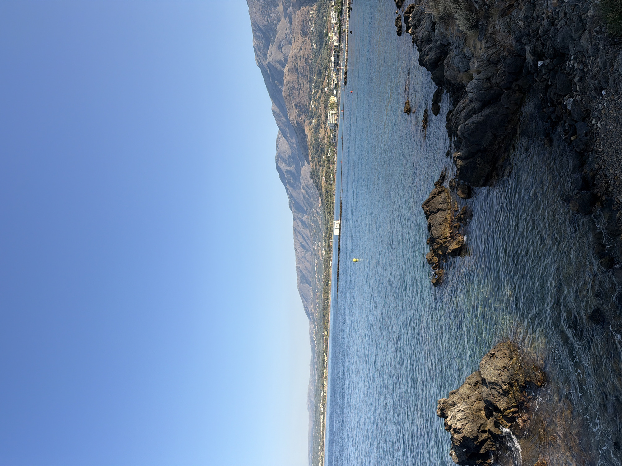 The image depicts a serene coastal scene in Georgioupoli, Greece, under a clear blue sky. The foreground is dominated by dark, weathered rocks that line the shore, with some partially submerged in the shallow, clear blue water, revealing the sandy seabed beneath. The water is calm, with gentle ripples lapping against the rocks.
In the mid-ground, the tranquil sea stretches out towards a distant coastline. A few buoys, including a prominent yellow one, are visible in the water. Further out, a town or village is nestled along the coast, with buildings and structures clustered together. Behind the town, a range of mountains rises, their slopes covered in dry vegetation and appearing in shades of brown and green. The overall impression is one of natural beauty and peacefulness. There are no people or animals visible in the image.