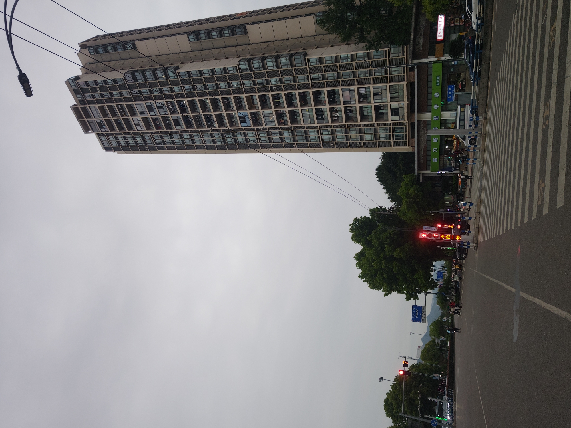 The image captures a street scene in what appears to be the Chongqing High-tech Zone, China. A tall, rectangular building with numerous windows dominates the left side of the frame, with multiple floors. In front of the building, there's a commercial establishment, possibly a store or center, indicated by signage that includes the characters "富力城里中心" and "富力城" (Fuli Chengli Center and Fuli Cheng, respectively), along with other Chinese text.
The right side of the image features a road with zebra crossings for pedestrians. Several traffic lights are visible, some showing red lights. Utility poles and power lines cross the sky. A sign that appears to display a time period, possibly related to traffic regulations, reads "710-9-30 17:00-19:00." The sky is overcast, and the overall environment seems urban, featuring street infrastructure, vehicles, and greenery along the roadside.