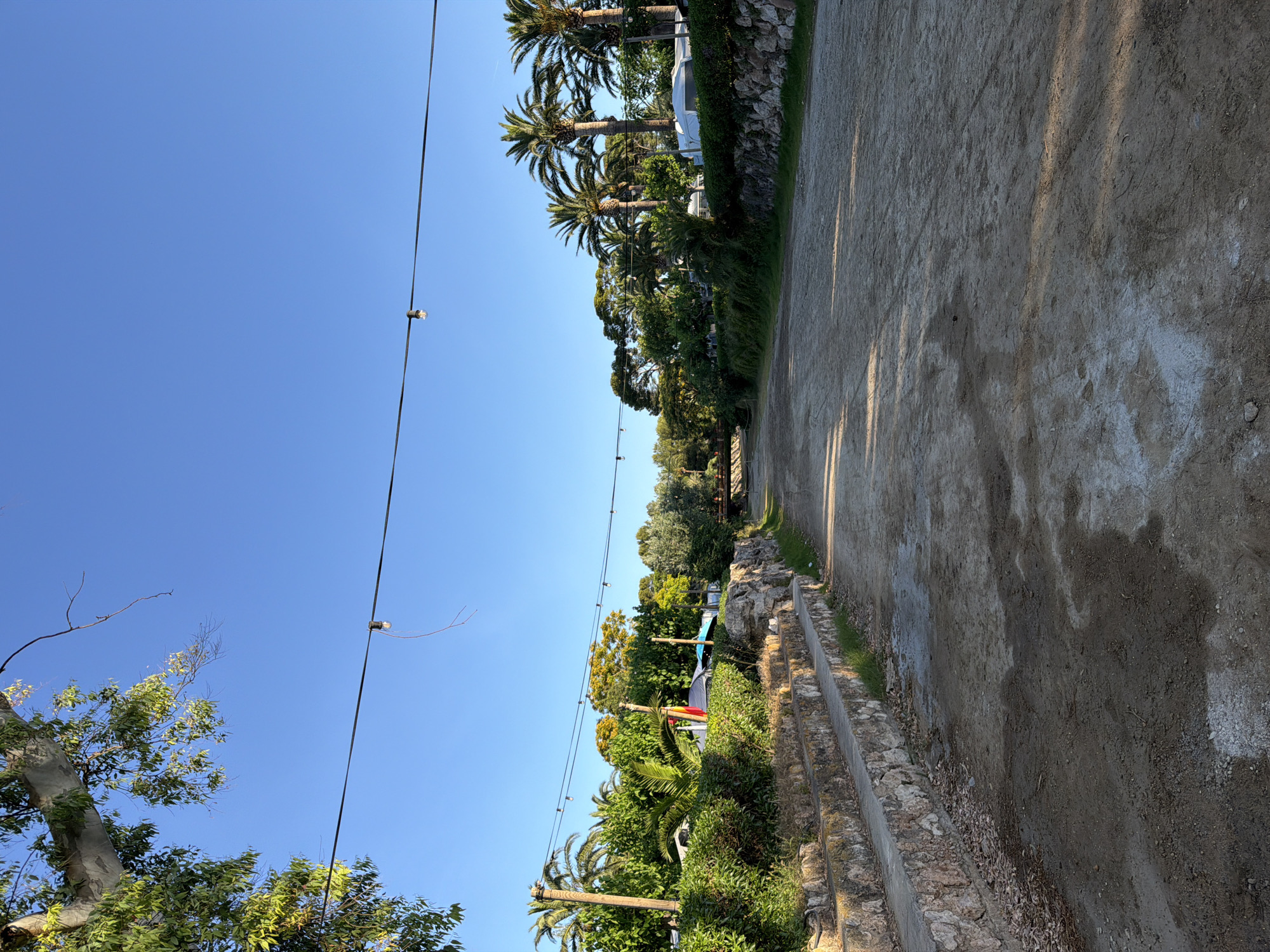 This image depicts a dry riverbed in Mont-roig del Camp, Spain. The riverbed is composed of dirt and sand, with some patches of dried leaves and white mineral deposits. On the left side of the riverbed, there are tiered stone steps leading up to a grassy area and lush vegetation. Beyond the vegetation, several tents and RVs are visible, suggesting a campsite. Palm trees and other leafy trees are scattered throughout the landscape, indicating a warm climate. A string of bare light bulbs hangs across the scene, strung between poles or trees. In the distance, a small bridge crosses the riverbed. The sky is clear and blue, with the sun casting shadows on the ground. There are no people visible in the image.