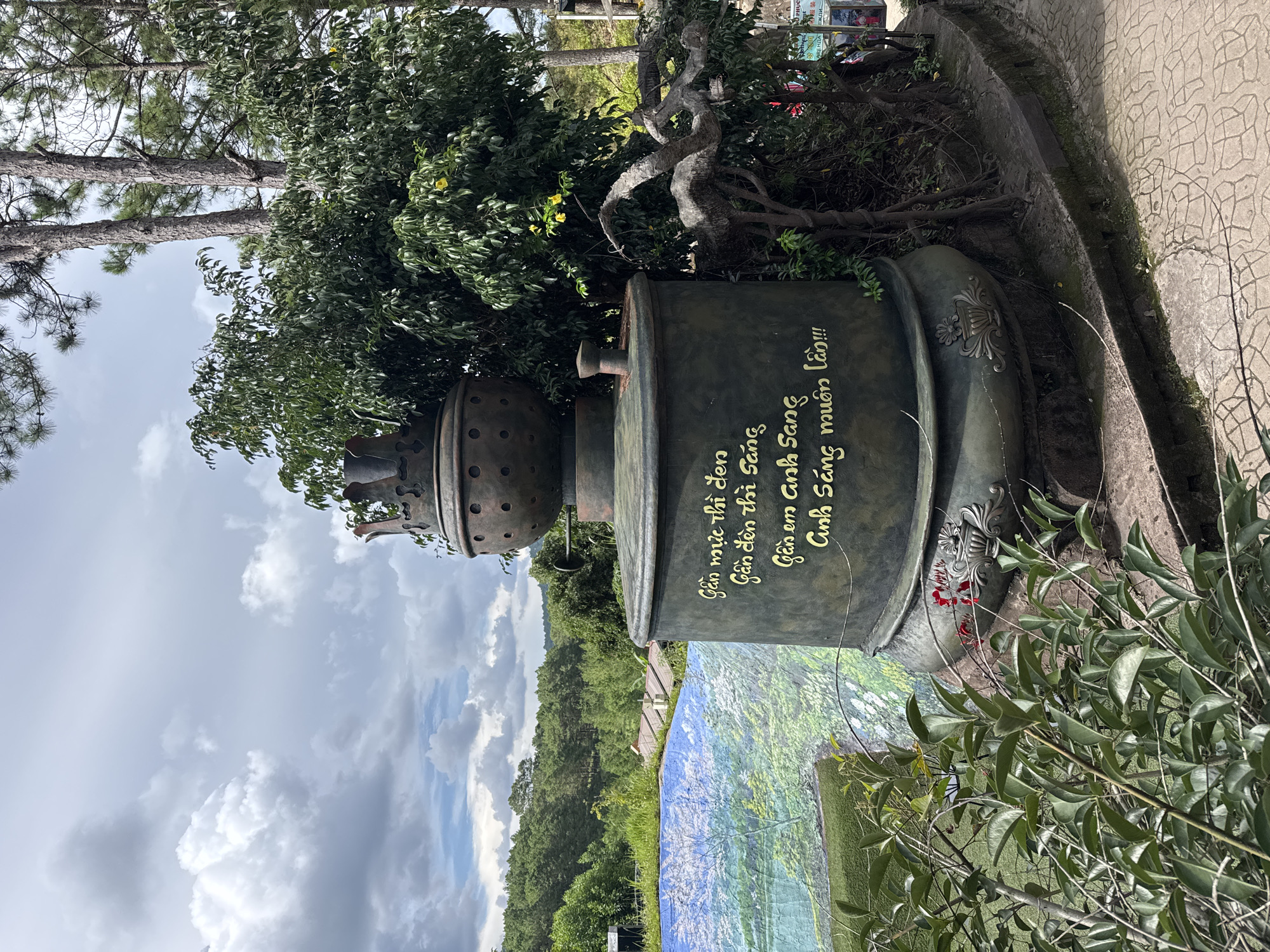 This is a vertical, outdoor photograph taken in Da Lat, Vietnam. The primary subject is a large, dark green, cylindrical object, possibly a sculpture or a piece of decorative equipment, with ornate gold lettering on its surface. The text, written in Vietnamese, reads: "Gần mực thì đen, Gần đèn thì sáng, Gần em anh Sang, Anh Sáng muôn lần!!!" which translates to "Close to ink, you are black. Close to light, you are bright. Close to you, I am Sang. I am bright a thousand times!". Above and around the object, lush green trees and foliage are visible, with pine trees dominating the background. The weather appears to be overcast, with dramatic grey clouds in the sky. The ground around the object is a textured, light-colored paving, and a vibrant, mural-like image of a landscape with trees and water is partially visible at the base of the cylinder. The overall impression is one of a decorative installation in a natural, possibly tourist, setting, with a touch of artistic expression through the inscribed text. There are no people visible in the frame. The lighting suggests it is daytime, but the overcast conditions may obscure direct sunlight. The prominent trees and the cool color palette of the sky hint at a mountain or highland region, consistent with Da Lat's climate.