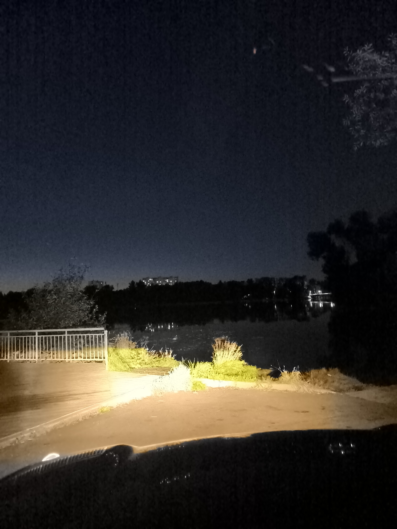 This image captures a nighttime view of a waterfront area in Dolgoprudny, Russia. The foreground is dominated by the hood of a vehicle, illuminated by its headlights, casting a bright yellow-orange glow on the pavement and a nearby wooden walkway. To the left of the walkway is a white metal railing. Beyond the walkway, the scene opens up to a calm body of water, reflecting the dim lights of distant buildings and trees that line the opposite shore. The sky is dark and clear, with a faint reddish object visible in the upper portion of the frame. The overall atmosphere is serene and quiet, suggesting a late-night or early morning setting.