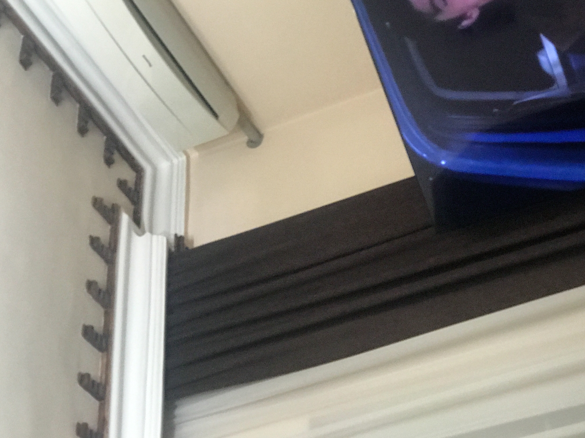 The image shows a partial view of an interior space, possibly a room, with a focus on architectural details and a partially visible electronic device. In the upper left corner, there's a section of decorative molding along the wall, with a distinct pattern of projecting elements. Adjacent to the molding, a white, rectangular object appears to be mounted on the wall, possibly an air conditioning unit or a similar appliance.
The lower portion of the image shows a window with blinds or shades. Dark, horizontal lines suggest the closed slats of the blinds. Overlapping the right side of the window is a fragment of a large, blue electronic device, potentially a television screen or a monitor. The screen displays a blurred image with pink and blue hues.
Based on a general understanding, it's impossible to identify with certainty that this scene is in Yerevan, Armenia. Additional information is necessary to confirm the geographical location.