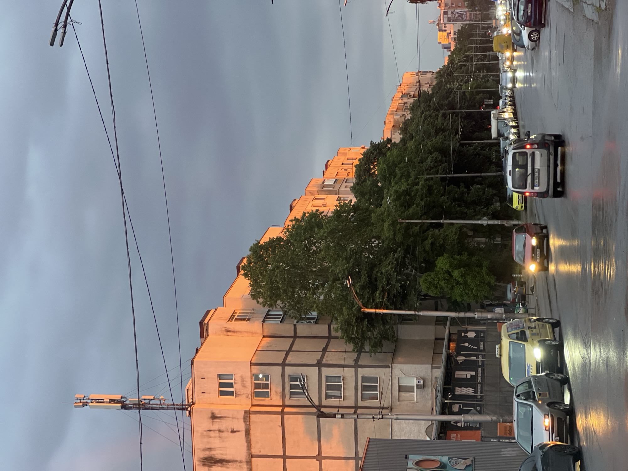 The image depicts a street scene in Sofia, Bulgaria, likely photographed after rain, given the wet asphalt. The foreground shows a dense row of parked and moving cars. In the middle ground, there is an apartment building illuminated by the setting sun or a similar warm light source. A line of trees borders the building. Overhead power lines are visible in the sky. The sky itself is overcast. An antenna array is attached to the side of a building.