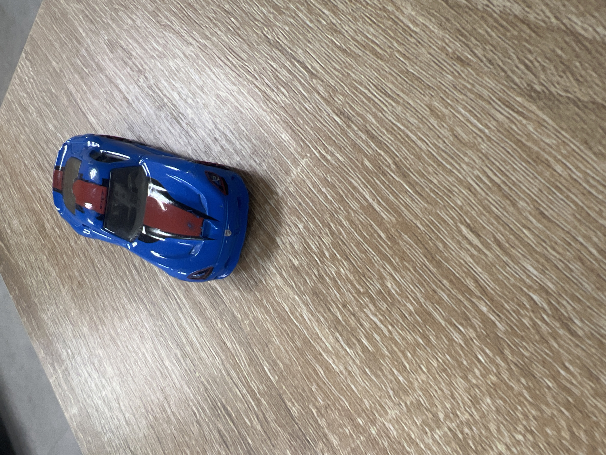 A blue and red toy car is positioned on a wooden surface. The car is facing towards the viewer and is slightly angled to the right. The wooden surface has a prominent grain pattern running diagonally from the top left to the bottom right. The background above the car appears to be a light gray surface, possibly a wall or another table. The image is an indoor shot, and there is no indication of the city of Minsk, Belarus, or any specific location within it. There are no people or any discernible actions occurring in the scene.