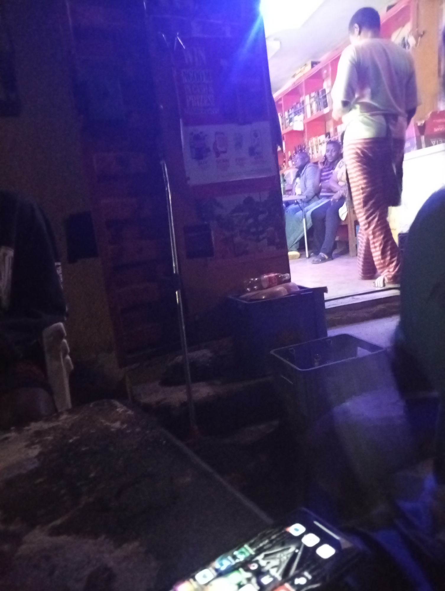The image, likely taken in Jos, Nigeria, shows a dimly lit indoor space, possibly a bar or small restaurant. A phone lies in the foreground, displaying a screen with various app icons. In the middle ground, plastic crates are stacked on steps. Behind them, there is a room with shelves stocked with bottles, presumably alcoholic beverages. Two people are sitting at a table, while another person in striped pants and light shirt stands with their back to the camera. A poster is partially visible on the wall, advertising winning prizes. The overall environment is dark and suggests a casual gathering place.
