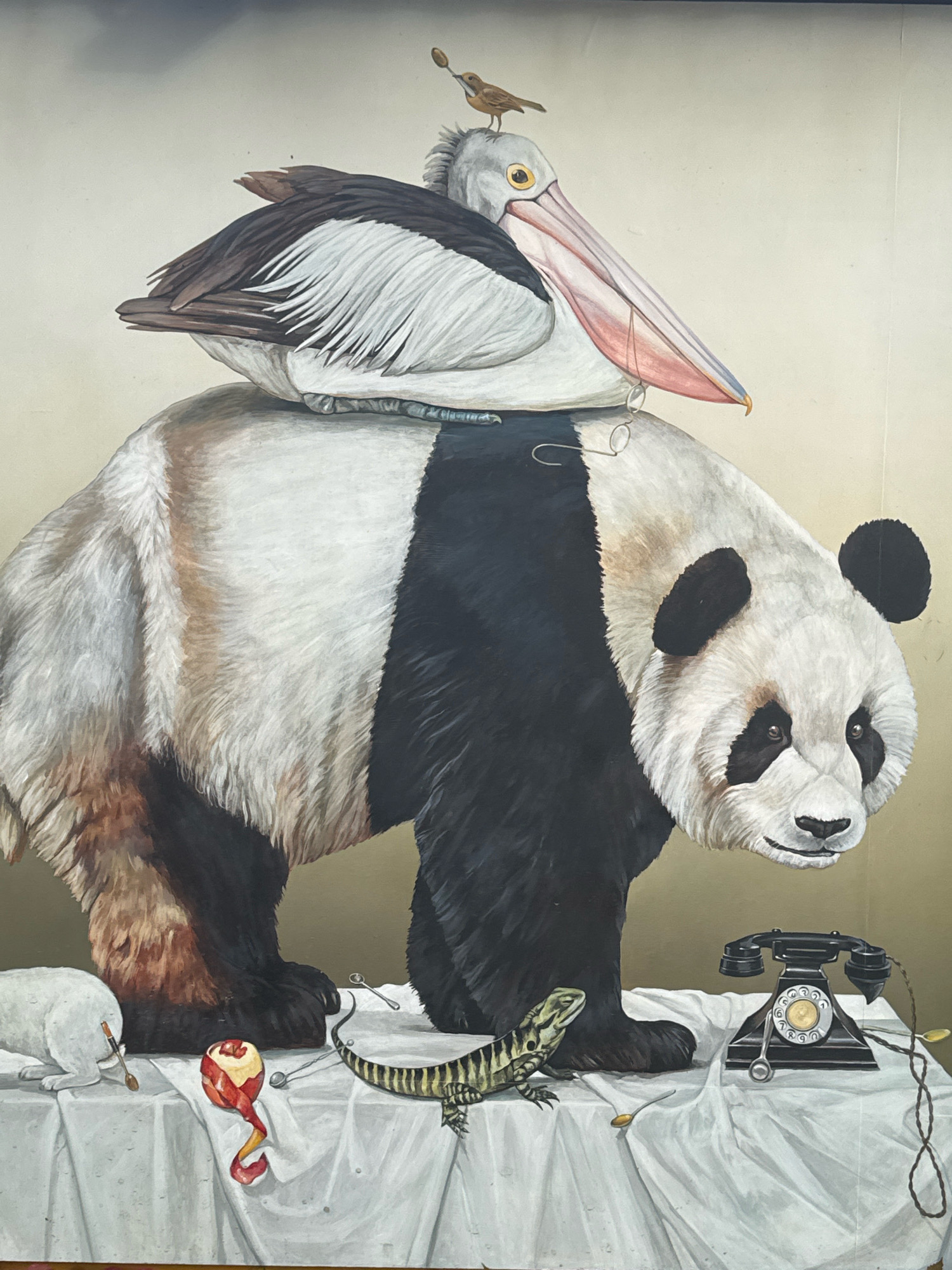 The image is a painting depicting a surreal scene. A large panda stands prominently in the center, with a pelican perched on its back. A tiny bird sits atop the pelican's head. The panda is covered in fur and has characteristic black and white markings. The pelican has a long beak and is painted with detailed feathers.
Below the panda, a white sheet is draped over a surface, resembling a table. On this surface, various objects are arranged. To the left, a white rabbit is partially visible, with a spoon near its hindquarters and a tube of lipstick. In the center, a peeled apple with its peel spiraled around it rests on the sheet, accompanied by a spoon. To the right of the apple, a lizard with distinctive stripes is positioned. Further to the right, a vintage black rotary telephone sits on the sheet, with its cord extending outwards. A spoon rests on the telephone's dial. The background of the painting is a muted, gradient color, transitioning from a lighter shade at the top to a darker shade at the bottom. The overall style of the painting is realistic, with meticulous attention to detail in the rendering of the animals and objects.
The location "Интурист Снт, Russia" appears to be a typographical error or a misunderstanding as it does not correspond to a known location. This image does not provide any visual cues to indicate its specific geographic location within a city or any recognizable landmarks.