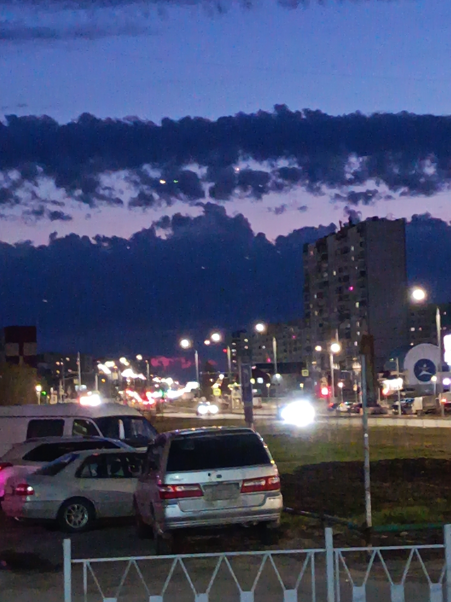 Captured at dusk, the image portrays a street scene in Nizhnevartovsk, Russia. The sky above is a blend of deep blue and violet hues, partially obscured by a layer of dark, textured clouds. Numerous streetlights dot the cityscape, their bright glows creating halos and reflections on the road. The scene includes automobiles, notably a white van, a silver sedan, and another silver vehicle in the foreground. There is a fence in the foreground. The cityscape is marked by tall buildings, suggesting a residential area, and a large, circular sign with an advertisement. The scene conveys an ordinary, nighttime atmosphere in the Russian city.