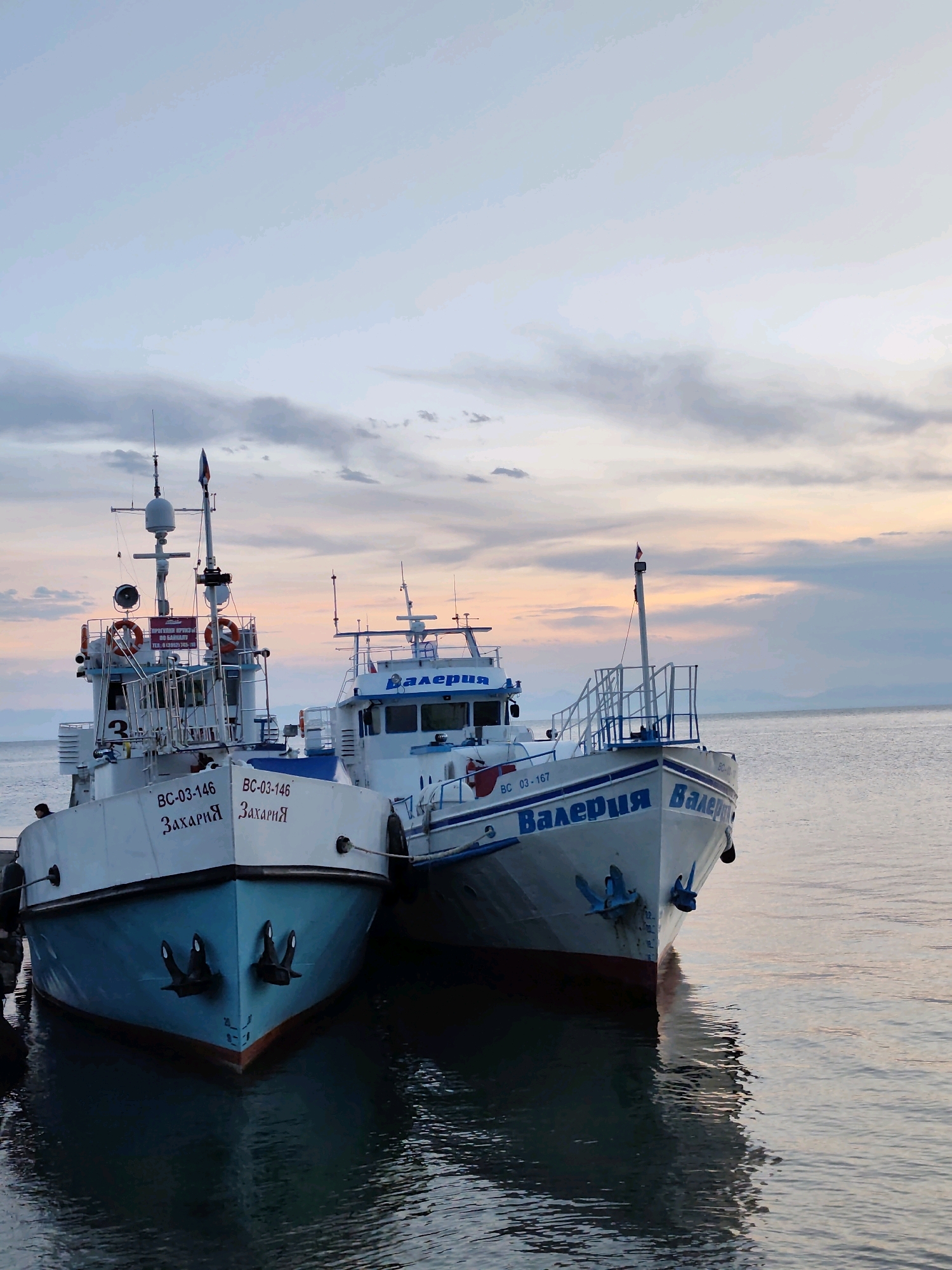 Two white boats with blue trim are docked side-by-side on calm water at dusk. The boat on the left is labeled "Захария" (Zacharia) in Cyrillic script, and the boat on the right is labeled "Валерия" (Valeria) in Cyrillic script. Both boats have masts with antennas and flags. The sky is a soft gradient of blue, pink, and orange, with scattered clouds. The reflections of the boats are visible on the water. The scene is likely a port in Listvyanka, Russia, a village located on the southwestern shore of Lake Baikal. A person is visible on the deck of the left boat, and another person is standing near the bow of the left boat. The overall atmosphere is peaceful and serene.