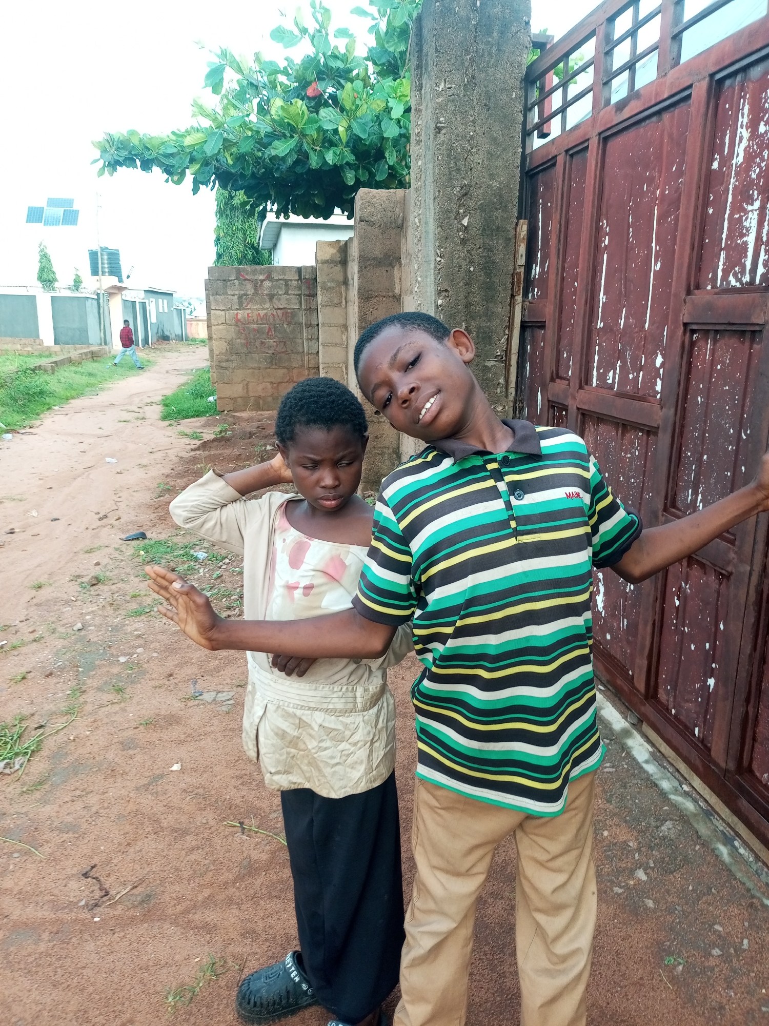 Two children, a boy and a girl, stand in front of a rusty, corrugated metal gate in what appears to be a residential area in Tempure, Nigeria. The boy, on the right, wears a striped polo shirt with green, yellow, and black horizontal bands and tan pants. He is leaning back with his head tilted and a slight smile. The girl, on the left, is wearing a tan, long-sleeved top over a patterned dress and black pants. Her left hand is raised, palm out, as if in a defensive or playful gesture, and she is looking downwards with a serious expression. The ground is dirt and gravel, with some scattered green grass. In the background, there is a stone wall, a tall tree, and buildings with solar panels. A person in a red shirt is visible walking in the distance.