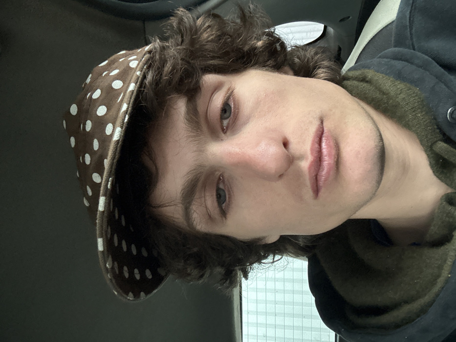 A close-up, frontal portrait of a male individual is captured within what appears to be the interior of a vehicle, situated in Vitry-sur-Seine, France. The subject, possessing medium-toned skin, dark wavy hair, and blue eyes, maintains a neutral facial expression while looking directly towards the camera. He wears a brown flat cap adorned with white polka dots, a dark-colored hooded outer jacket, and an olive-green hooded garment beneath. The background on the left reveals a window through which a light-colored structure with a prominent grid or horizontal line pattern is visible. To the right, a dark, indistinct vehicle interior component is partially discernible. The lighting is even, suggesting an enclosed space with diffused external light.
