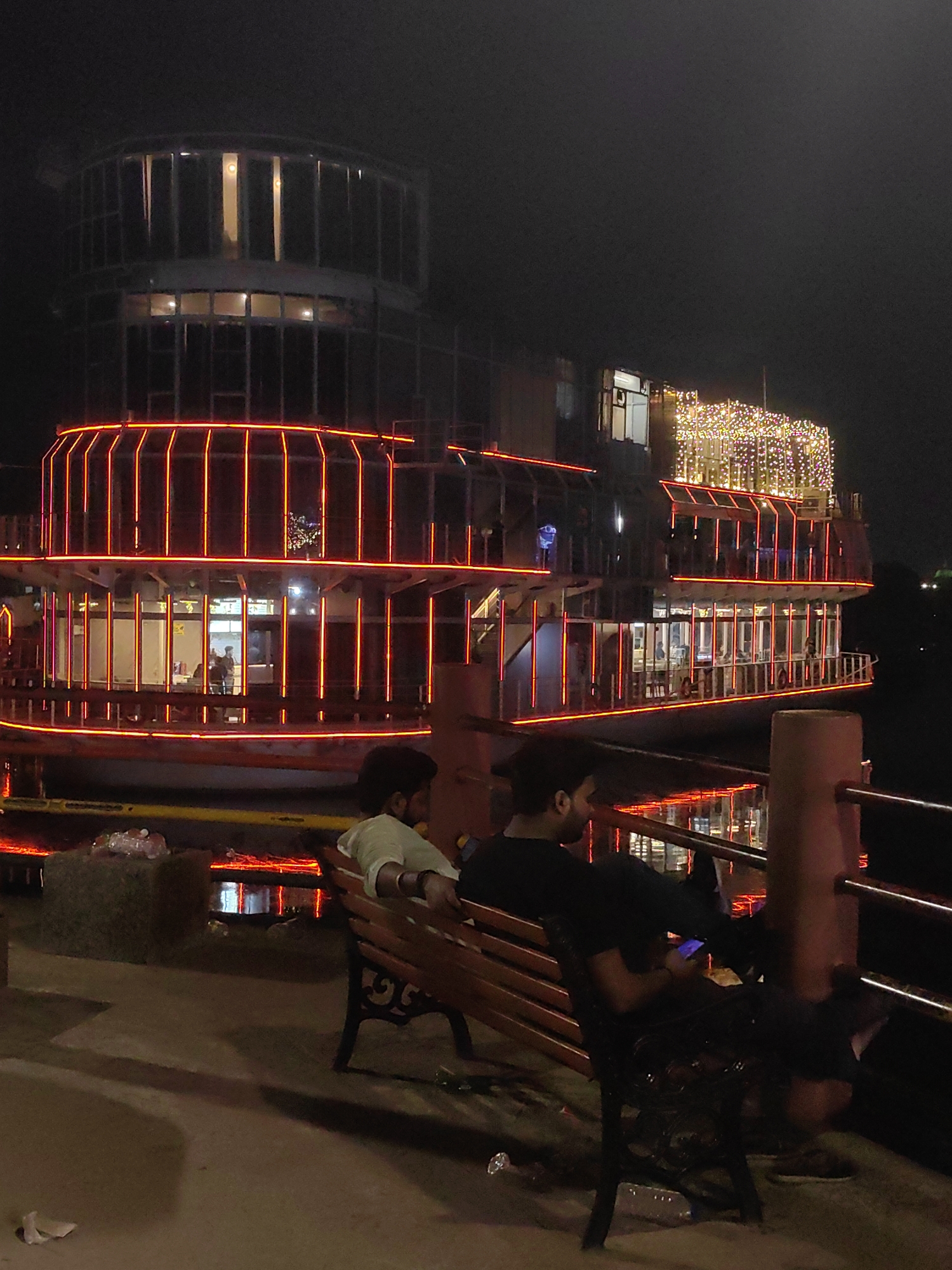 In Gorakhpur, India, at night, two individuals are seated on a wooden bench facing away from the camera, towards a large, illuminated boat docked on a body of water. The boat has multiple levels and is adorned with bright red and yellow lights. One person, on the left, wears a light-colored shirt and is looking forward. The other person, on the right, wears a dark shirt and is looking down at a mobile phone. The bench is positioned on a paved walkway or embankment, with a metal railing in the foreground. The environment appears to be a public space, possibly a waterfront promenade, with subtle reflections of the boat's lights visible on the water's surface.