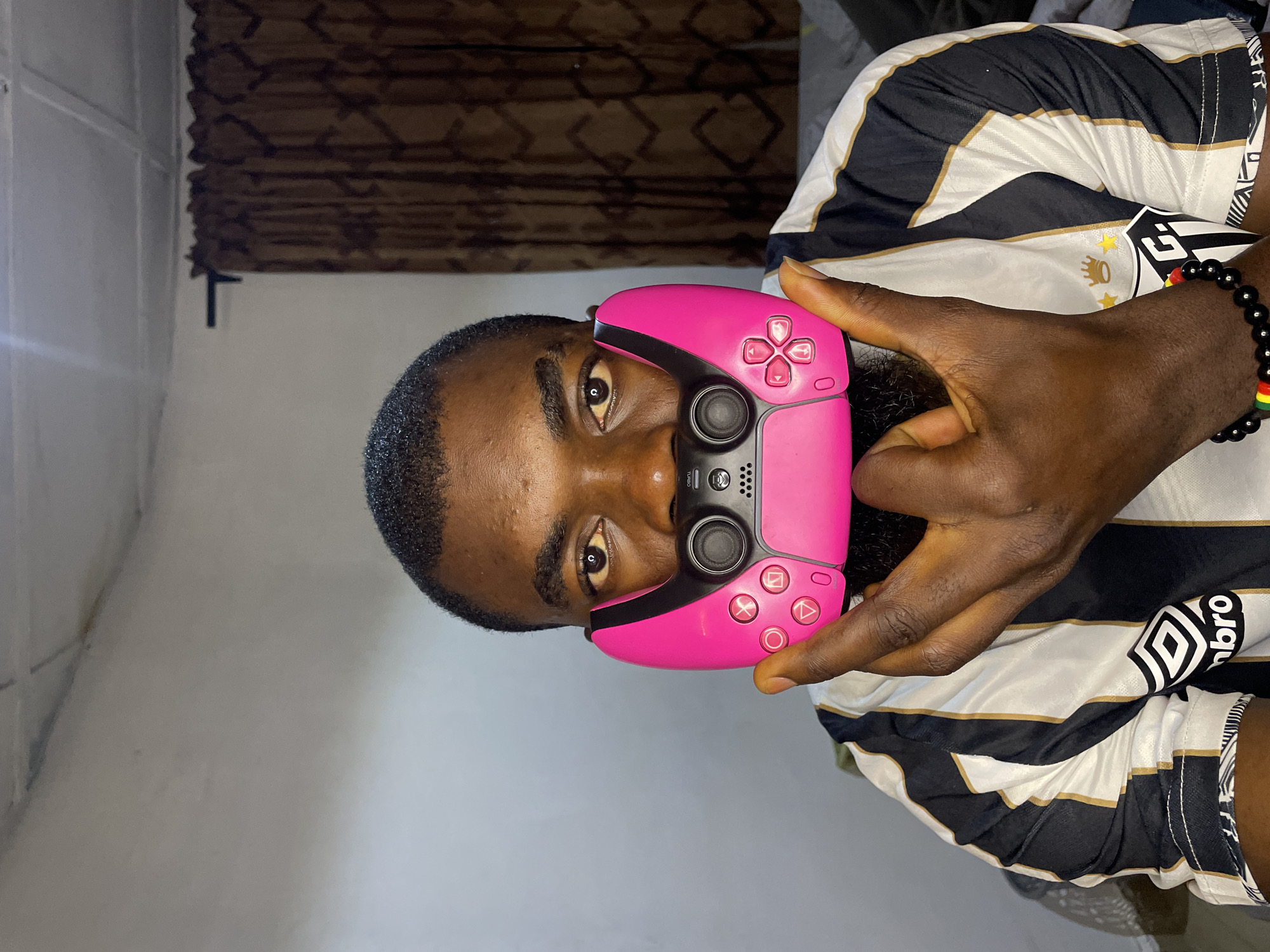 In Jos, Nigeria, a man is pictured from the chest up, holding a bright pink video game controller to his face as a makeshift mask. He is wearing a black and white striped jersey with gold trim, and a bracelet with red, yellow, and green beads. His left hand is raised, with his thumb and index finger extended. The background consists of a plain white wall and a brown patterned curtain.