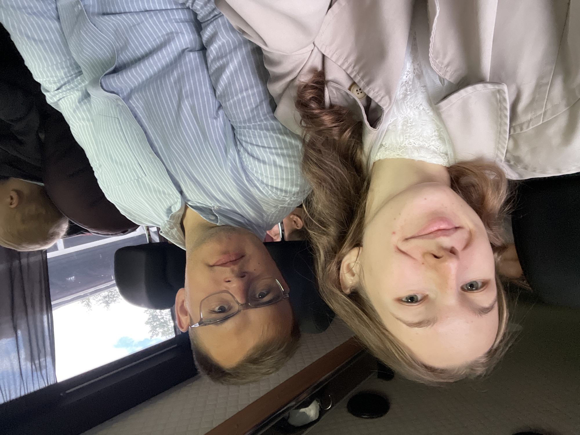 The image shows a close-up, waist-up, selfie-style shot of two people, a woman and a man, sitting in what appears to be the interior of a vehicle, possibly a bus or a van. The woman is on the left, her head tilted slightly and her eyes looking towards the camera with a slight smile. She has blonde hair that falls around her shoulders and is wearing a light-colored trench coat over a white lace top. The man is on the right, wearing glasses and a light blue and white striped button-down shirt. He is looking directly at the camera with a neutral expression. In the background, to the right of the man, a small child with blonde hair is visible, leaning back against a seat. The interior of the vehicle shows dark upholstery on the seats and a glimpse of a window with dark curtains, suggesting it could be a mode of transport. The lighting is somewhat diffused, indicating an indoor setting or an overcast day. There is no specific indication of the location being the city of Ulmen, Germany; the context is solely derived from the visual elements within the image.