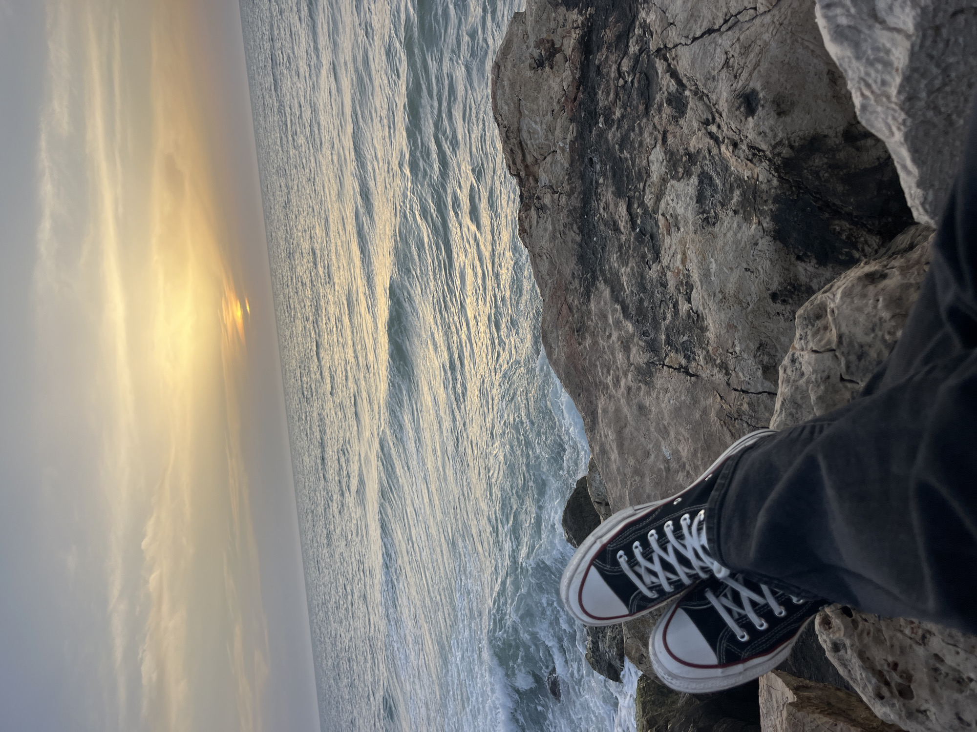 The image shows a view from a rocky cliff overlooking the Mediterranean Sea at sunset. In the foreground are a pair of Converse-style sneakers and the leg of the person wearing them. The cliffs are dark gray and tan in color, with the sea stretching out towards the horizon where the sun is setting. The location is potentially along the coastline of Tel-Aviv, Israel. The photograph appears to be taken from a high vantage point, with the ocean and sky filling the majority of the frame.