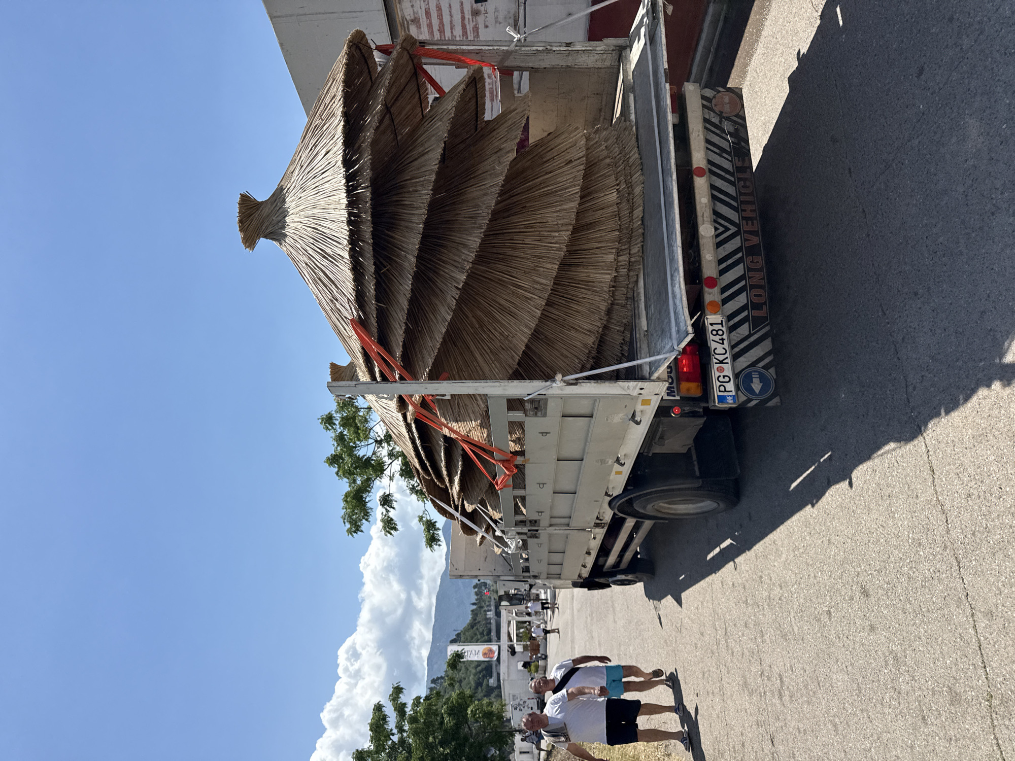 The image, taken in Budva, Montenegro, depicts a white flatbed truck transporting numerous large, thatched beach umbrellas (palapas) on a sunny day. The truck is positioned on a paved road, with its rear facing slightly towards the viewer. The circular, straw or reed-covered umbrellas are stacked horizontally in the truck bed, secured with bright red straps, showcasing their rustic texture and distinctive pointed tops.
On the paved road to the right of the truck, two men are walking away from the vehicle. One wears a white T-shirt and light blue shorts, while the other is in a white T-shirt and dark shorts. Their distinct, sharp shadows on the asphalt indicate bright, overhead sunlight.
The background features a clear blue sky with some large, white clouds hovering over a range of green, tree-covered mountains. Some distant buildings and additional trees are also visible, suggesting a vibrant outdoor setting.
Visible text on the truck's rear includes a "LONG VEHICLE" sign with diagonal black and white stripes. Below this, a Montenegrin license plate, "PG KC481," is clearly legible, with "PG" being the code for Podgorica. The scene captures an everyday activity of transporting seasonal items, likely to or from a beach resort, under clear skies and warm weather.