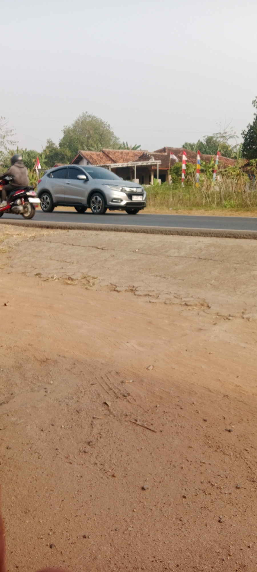 A silver Honda HR-V is driving on a paved road in what appears to be a rural area. To the left of the car, a person on a red and black motorcycle is also traveling on the road. In the background, there are several houses with terracotta roofs, surrounded by vegetation and trees. Several Indonesian flags are also visible in front of the houses. The foreground of the image is a dirt or sand-covered area. The sky is overcast and pale.