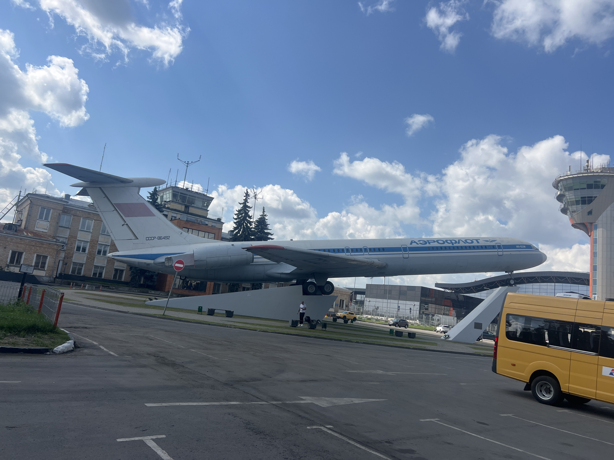 An outdoor, slightly angled, eye-level, long shot captures a sunny, partly cloudy day scene featuring a static display of an Aeroflot aircraft. The plane, marked with "CCCP-86492" and the Aeroflot logo, is mounted on a white, sloped pedestal. To the left of the plane, a brick building with numerous windows stands under a blue sky with white, puffy clouds. Behind the plane, a modern, glass-enclosed control tower with a rounded top is visible. In the foreground, a yellow minibus is parked at an angle, partially obscuring a paved area marked with white lines, indicating a parking lot or roadway. A person is standing near the base of the aircraft. The scene is set in the city of Khimki, Russia, identifiable by the architectural style of the buildings and the presence of a prominent Aeroflot aircraft, likely at an airport or aviation museum.