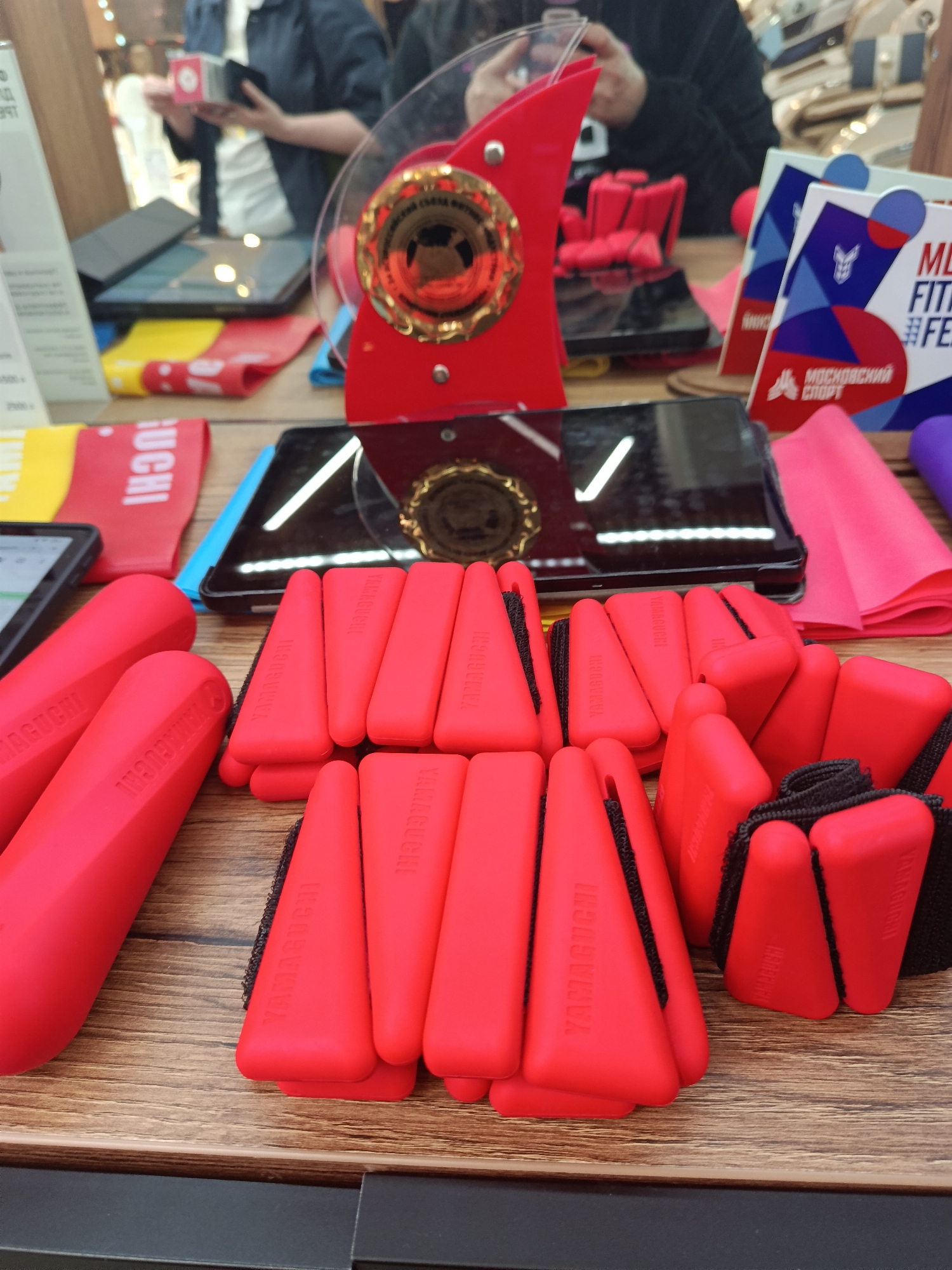 The image depicts a display in what appears to be a retail or promotional setting in Moscow, Russia. The text "МОСКОВСКИЙ СПОРТ" is visible. On a wooden surface, several fitness-related items are arranged. There are red, cylindrical hand weights with the name "YAMAGUCHI" printed on them, as well as smaller red triangular pieces also labeled "YAMAGUCHI" and connected by black straps. A black, rectangular device (likely a phone or tablet) sits behind these items, reflecting a gold-colored object (possibly a medallion or logo). Behind the device, there is a clear, curved stand with a red emblem and what appears to be a gold medallion at its center. In the background, there are more items and people examining the display. One person is holding a small, white box. Additional display materials in yellow, red, pink, and purple colors are scattered around.