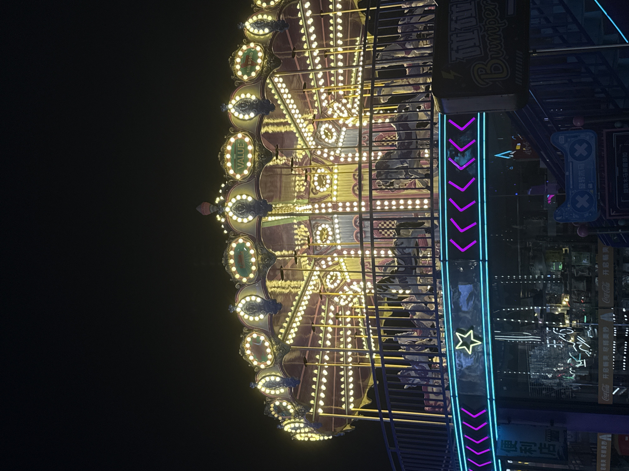 This is a night-time photograph of a brightly lit carousel in Yiwu, China, specifically at the Futian Market, which is a renowned international trade city. The carousel is adorned with numerous yellow lights along its roof and decorative elements. The carousel itself appears to be made of a red fabric with ornate golden trim. White horses, some with purple details, are visible on the carousel platform, suggesting it is operational or ready for operation.
Below the carousel, a railing with blue and purple neon lights forms a decorative border. A yellow star shape is illuminated within this neon border. In the background, reflections of lights suggest other attractions or shops, including what appears to be a convenience store with Chinese characters and Coca-Cola branding. To the right of the carousel, a dark sign with yellow lettering that reads "Bumper" is visible, indicating the presence of a bumper car attraction. The overall environment is dark, with the primary light sources being the artificial lights of the carousel and surrounding attractions. No people are clearly visible in the frame.