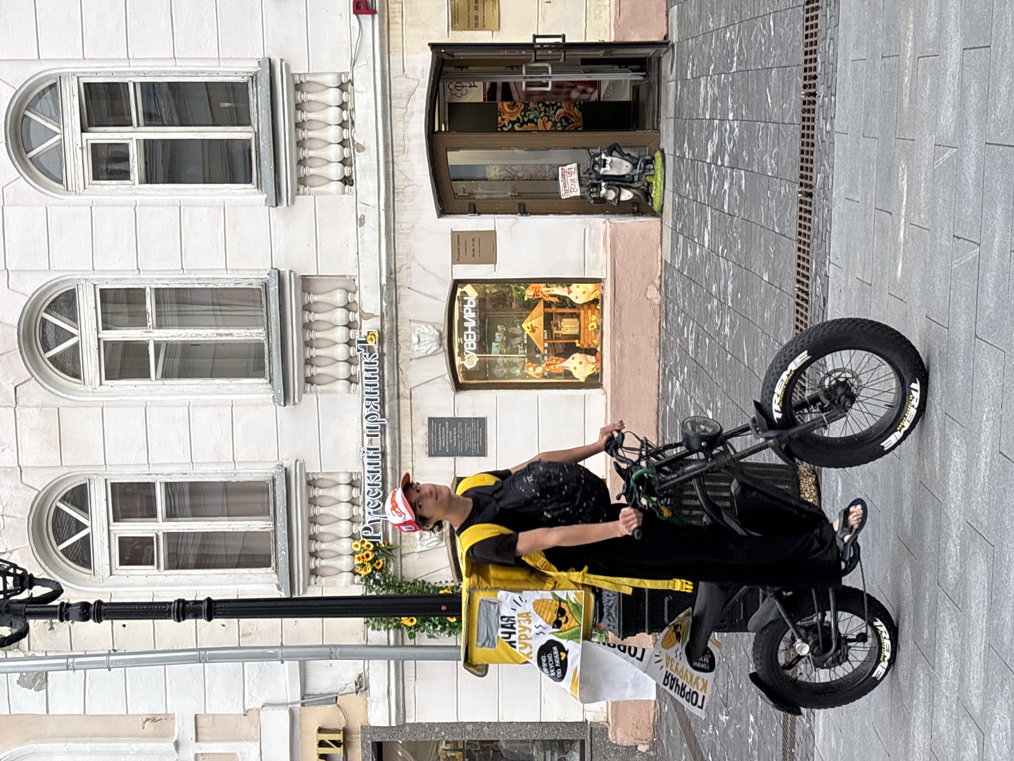 A young man is riding a black electric bicycle with wide tires on a paved street in front of a building with arched windows and ornate balconies. He is wearing a black t-shirt, black pants, flip-flops, and a red and white baseball cap. He has a black backpack on and a large yellow insulated delivery bag strapped to the back of the bicycle. A sign on the delivery bag features a cartoon corn character wearing sunglasses. Another sign, partially obscured, hangs from the handlebars and also features the corn character and Russian text. In the background, the building has several windows, a doorway leading into a shop, and decorative elements. A lamppost stands to the left of the man. The shops visible are identified by signs indicating "Русский Пряник" (Russian Gingerbread) and "Сувениры" (Souvenirs). The architecture suggests a historic urban setting. The context of "Русский Пряник" and the style of the building indicate this scene is likely in Nizhny Novgorod, Russia.