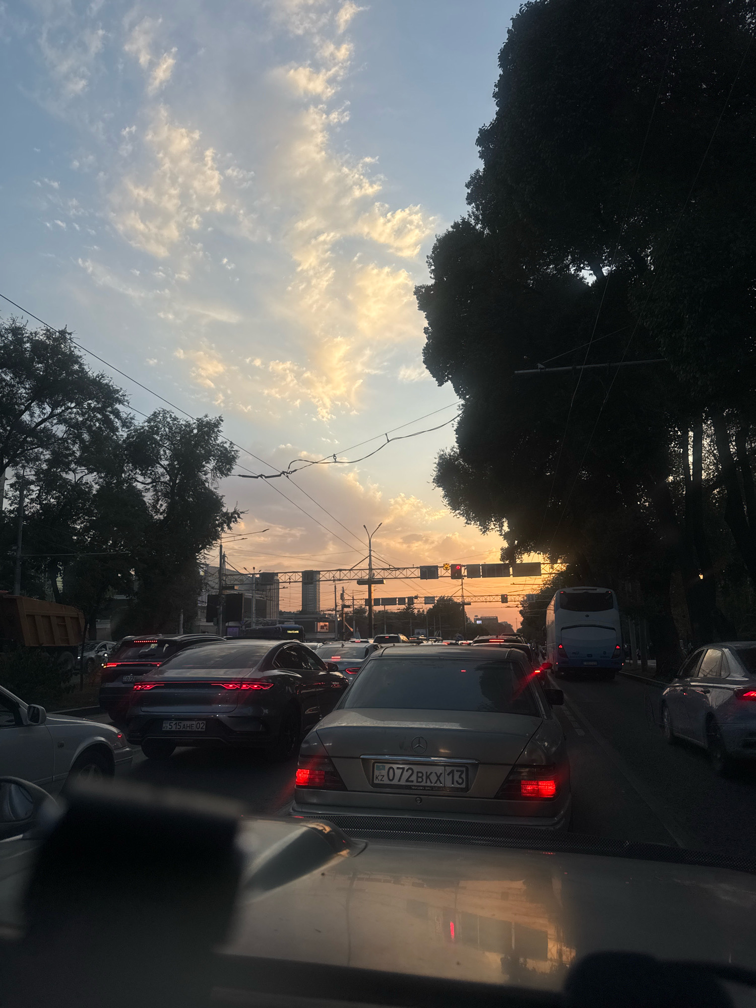 The image depicts a traffic jam on a city street in Almaty, Kazakhstan, during sunset. The sky is filled with dramatic clouds illuminated by the setting sun, casting a warm glow. Several cars are visible on the road, including a Mercedes-Benz with license plate "072 BKX 13" and a dark grey sedan with license plate "515 AHE 02." A blue bus is also present in the traffic. Trees line the right side of the street, casting shadows. Traffic lights and overhead power lines are also visible, indicating an urban environment. The overall scene suggests a typical evening commute with heavy traffic.